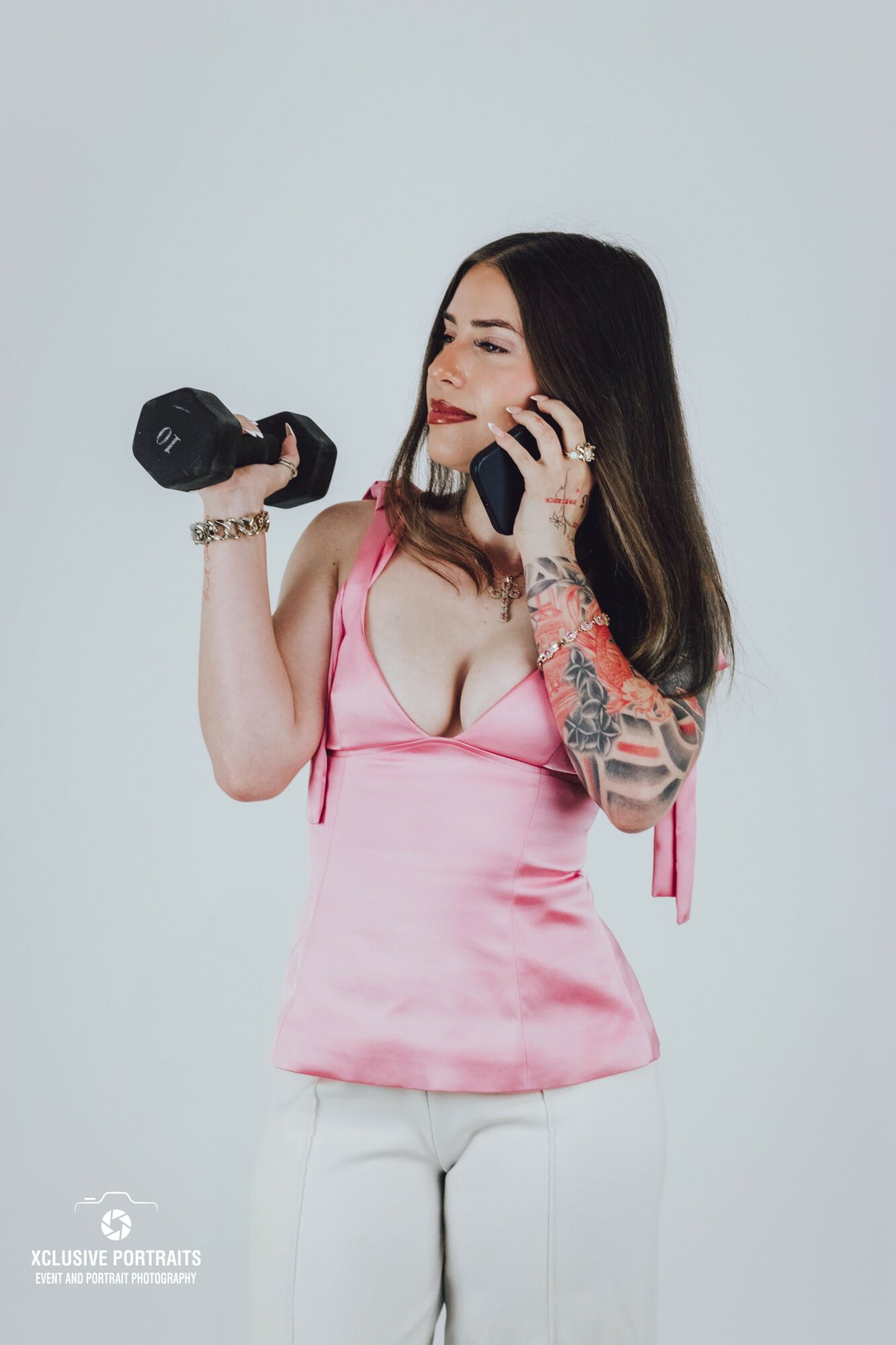 Woman with long dark hair in pink top, holding a dumbbell in one hand and a phone to her ear, against a plain background.