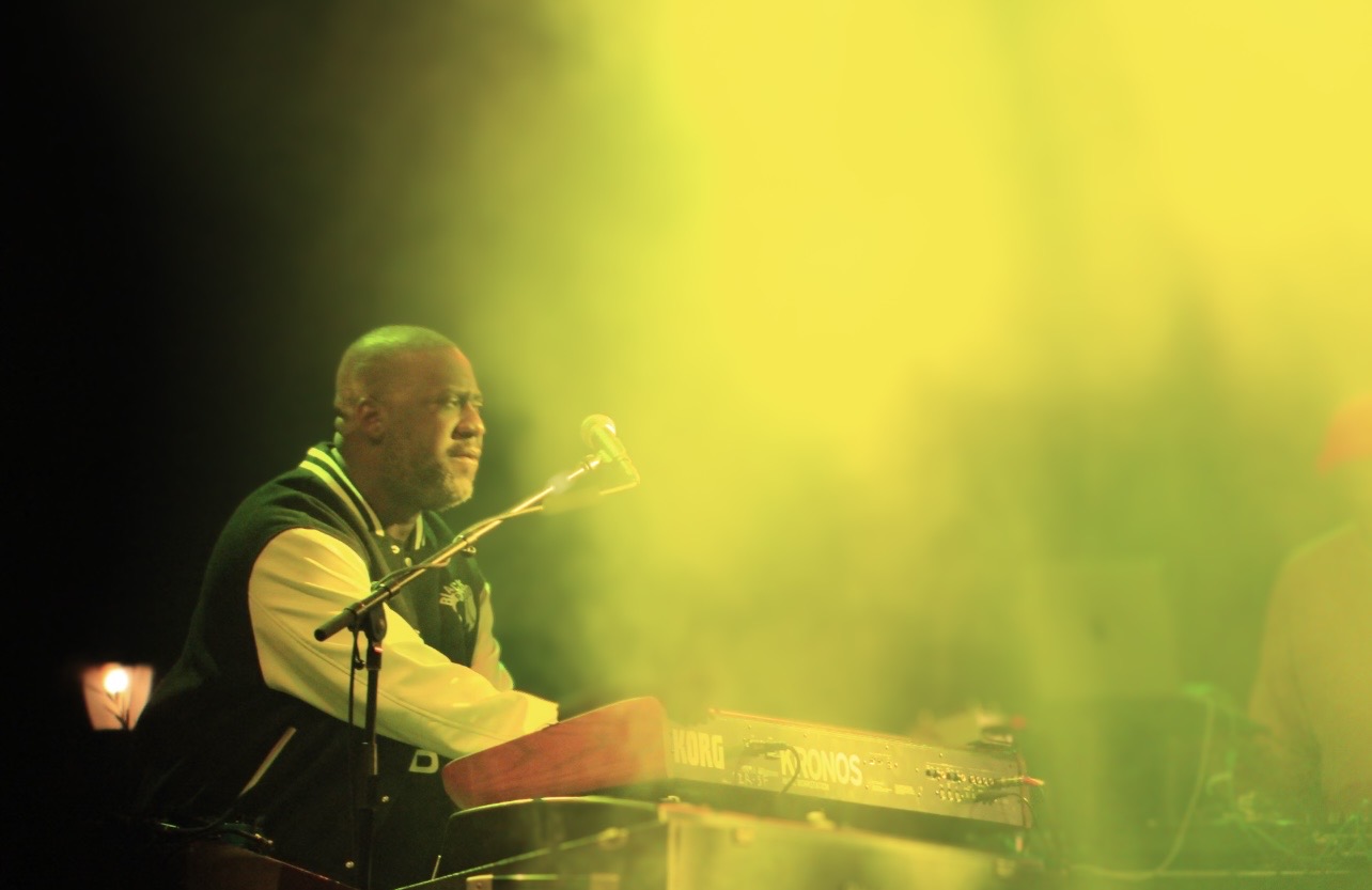 Person playing keyboard on stage with yellow lighting, microphone nearby, and a dark background.