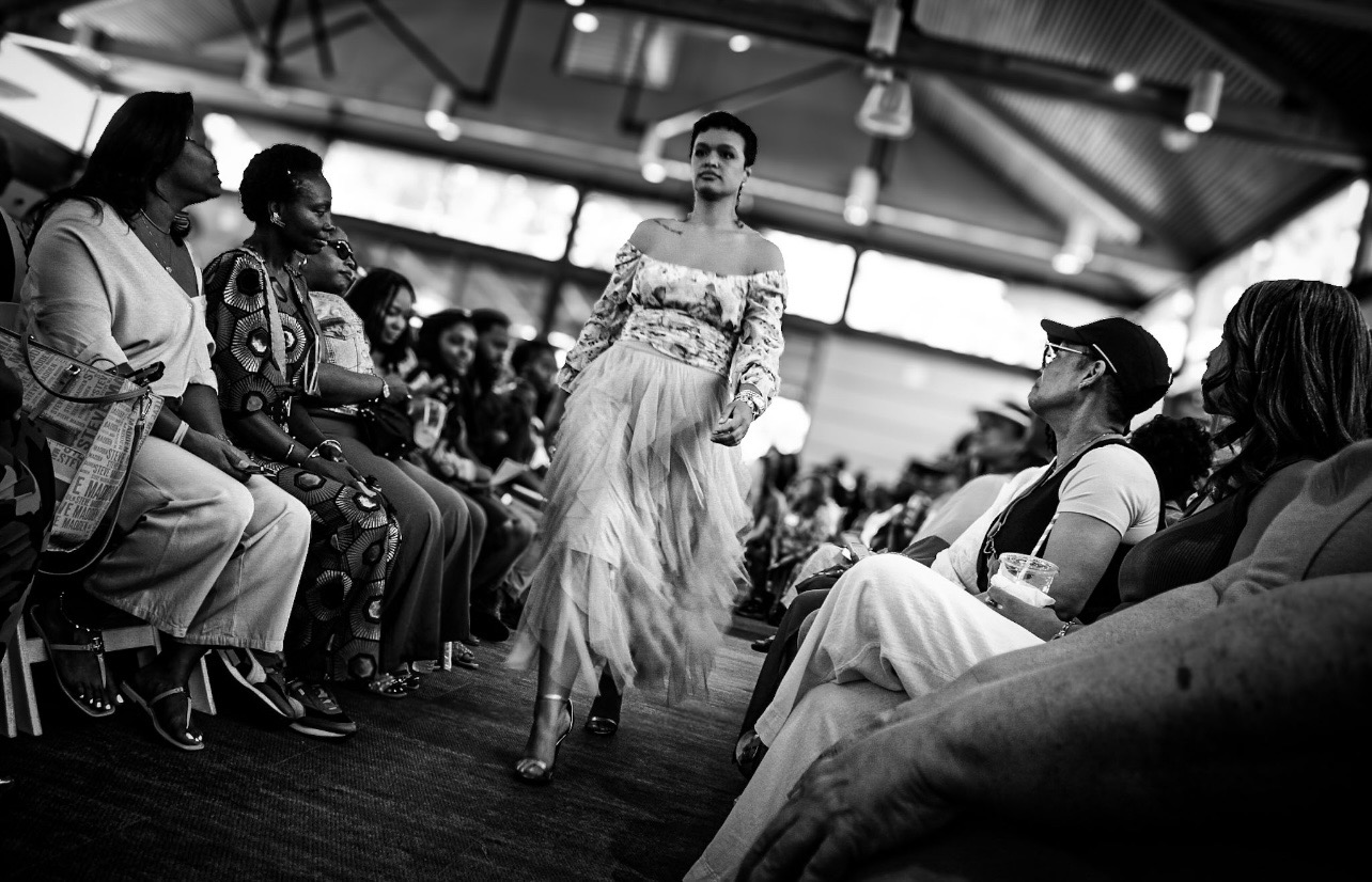 A woman walks on a runway in front of seated audience members in an indoor setting, with a high ceiling and large windows.