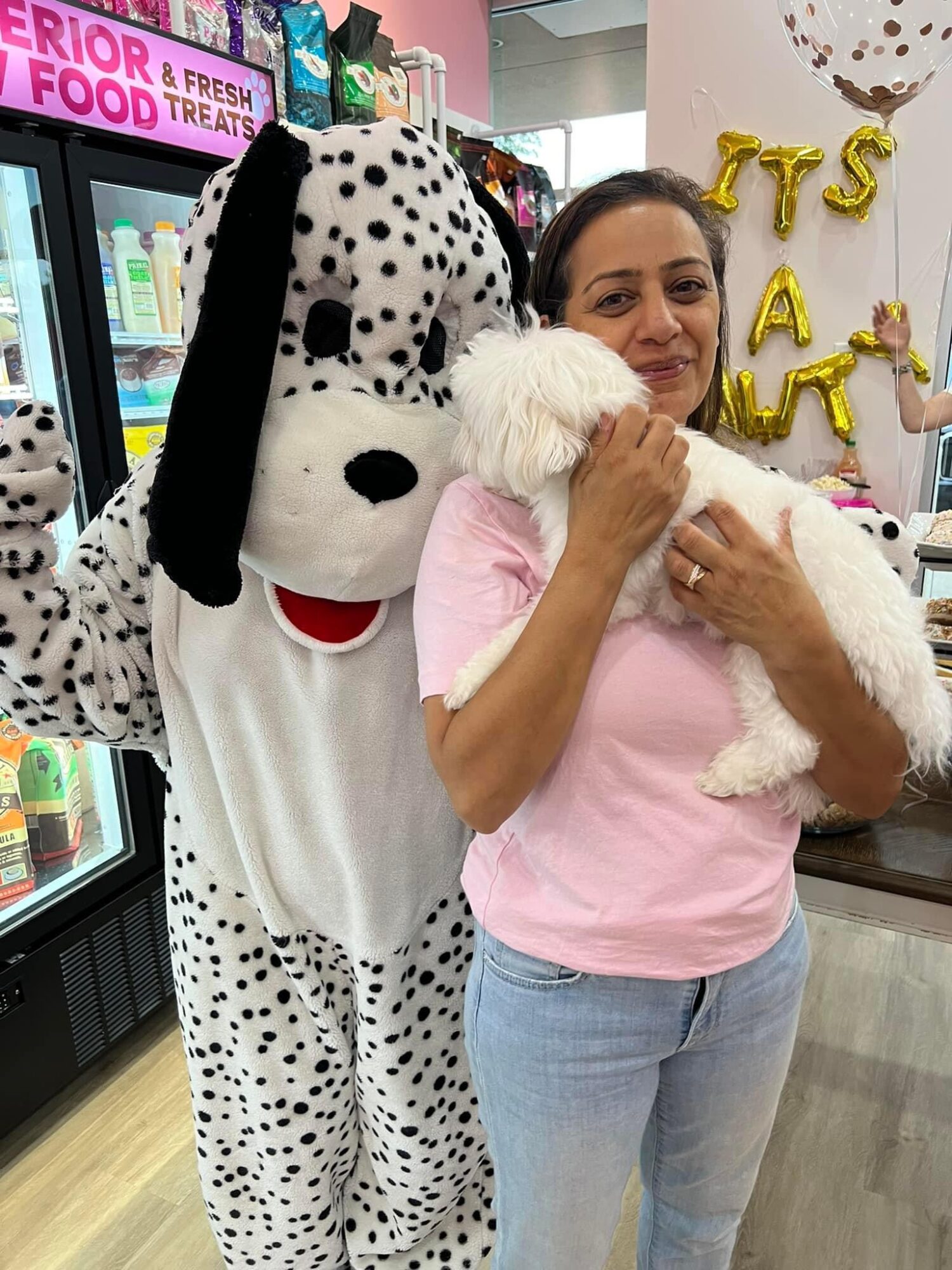 Woman holding a white puppy in a store, mascot dressed as Dalmatian nearby, shelves with products in background.