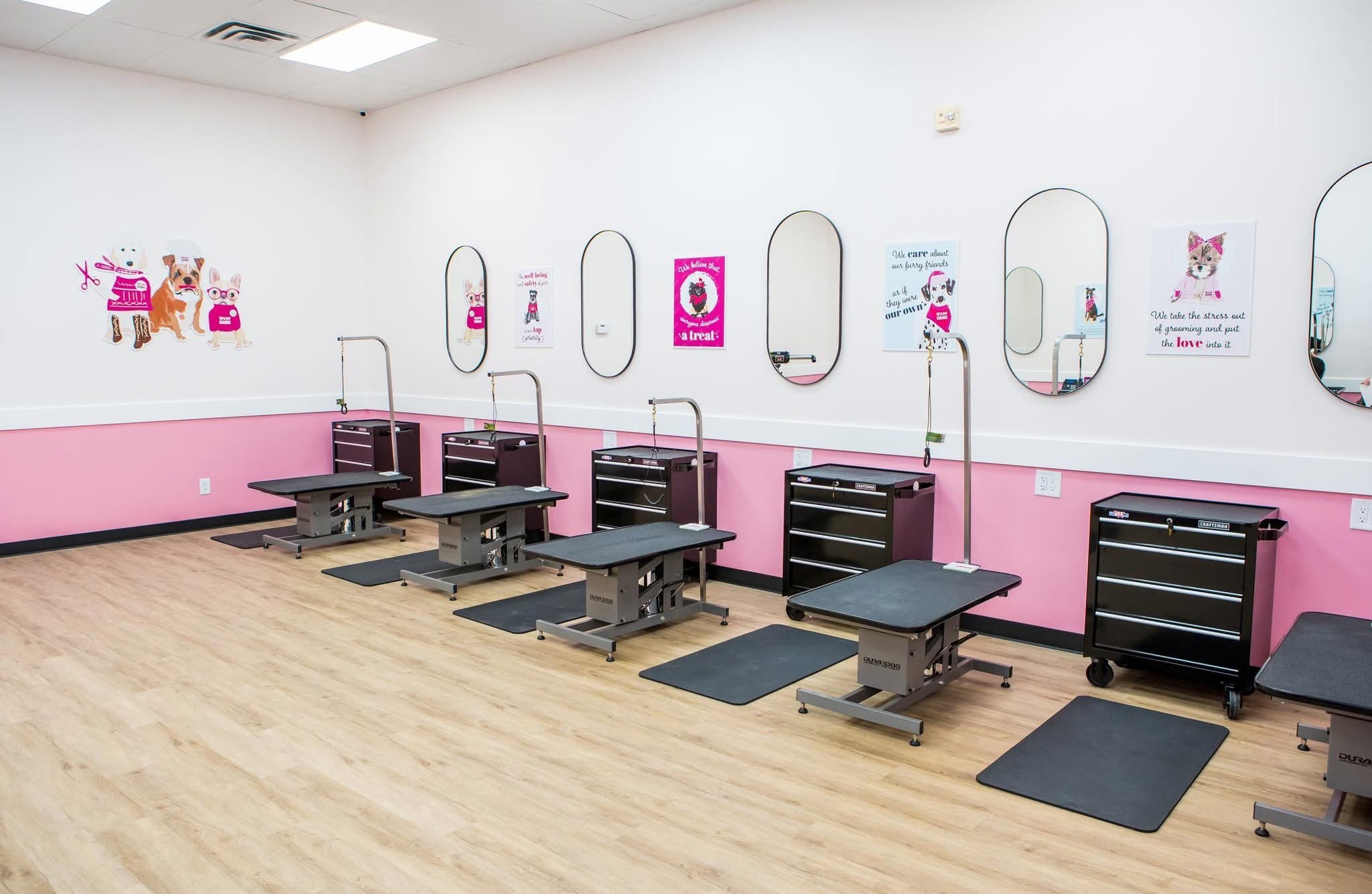 Room with five adjustable chairs, mirrors, and pink wall decorations, likely a beauty or hair salon, with wooden floor and black mats.