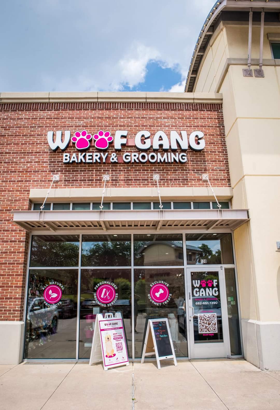 Storefront with large glass windows and a brick facade, signage for bakery and grooming business, sidewalk outside, sky visible above.