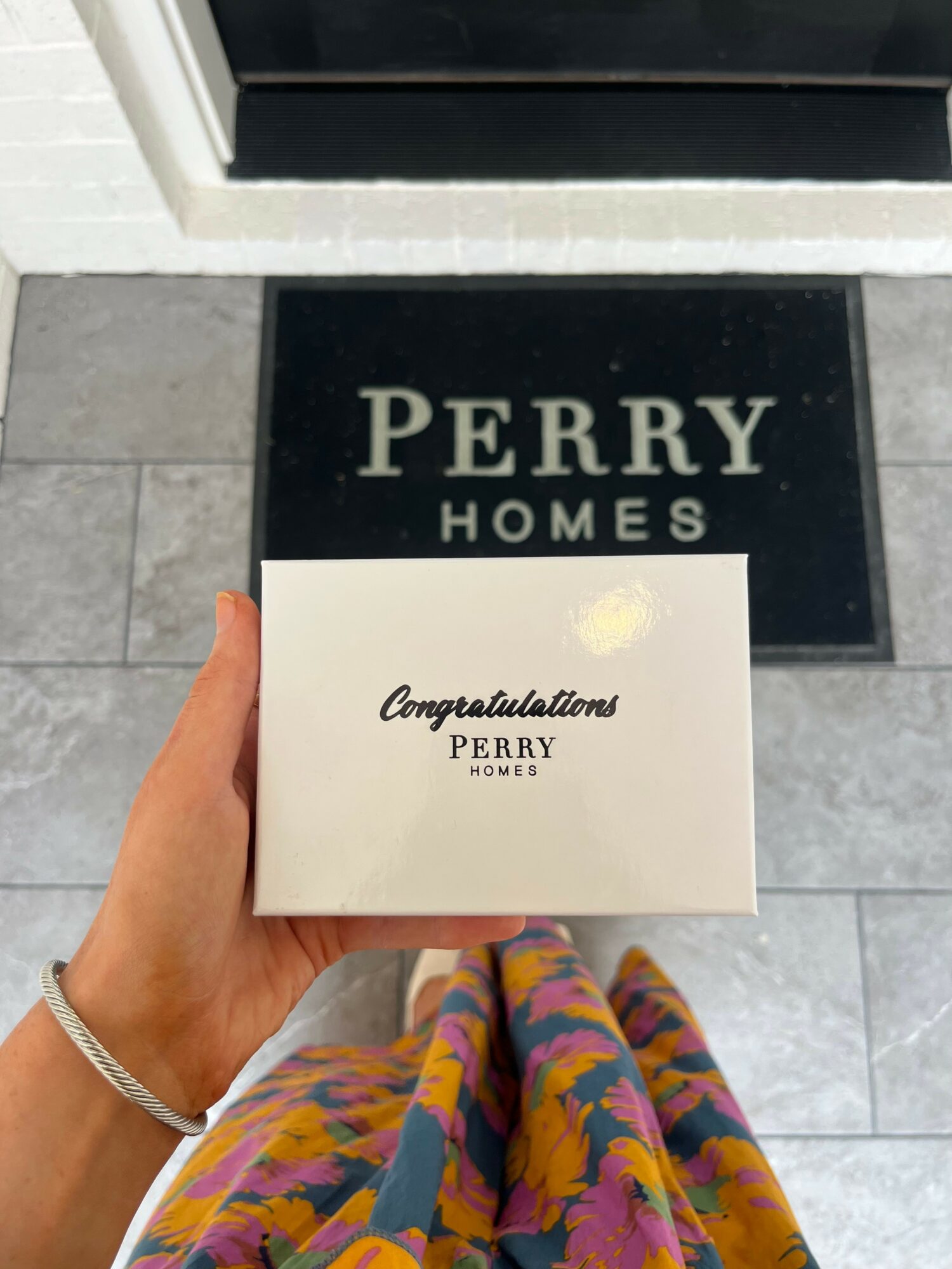 Hand holding a white box with congratulatory message in front of Perry Homes sign, with colorful skirt visible.