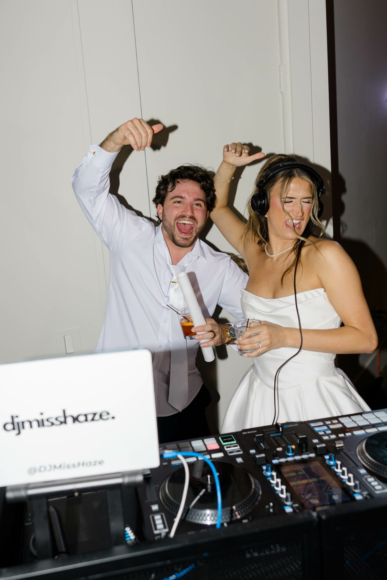 Two people, a man and a woman, are DJing and dancing at a party, smiling and holding drinks, with DJ equipment in front.