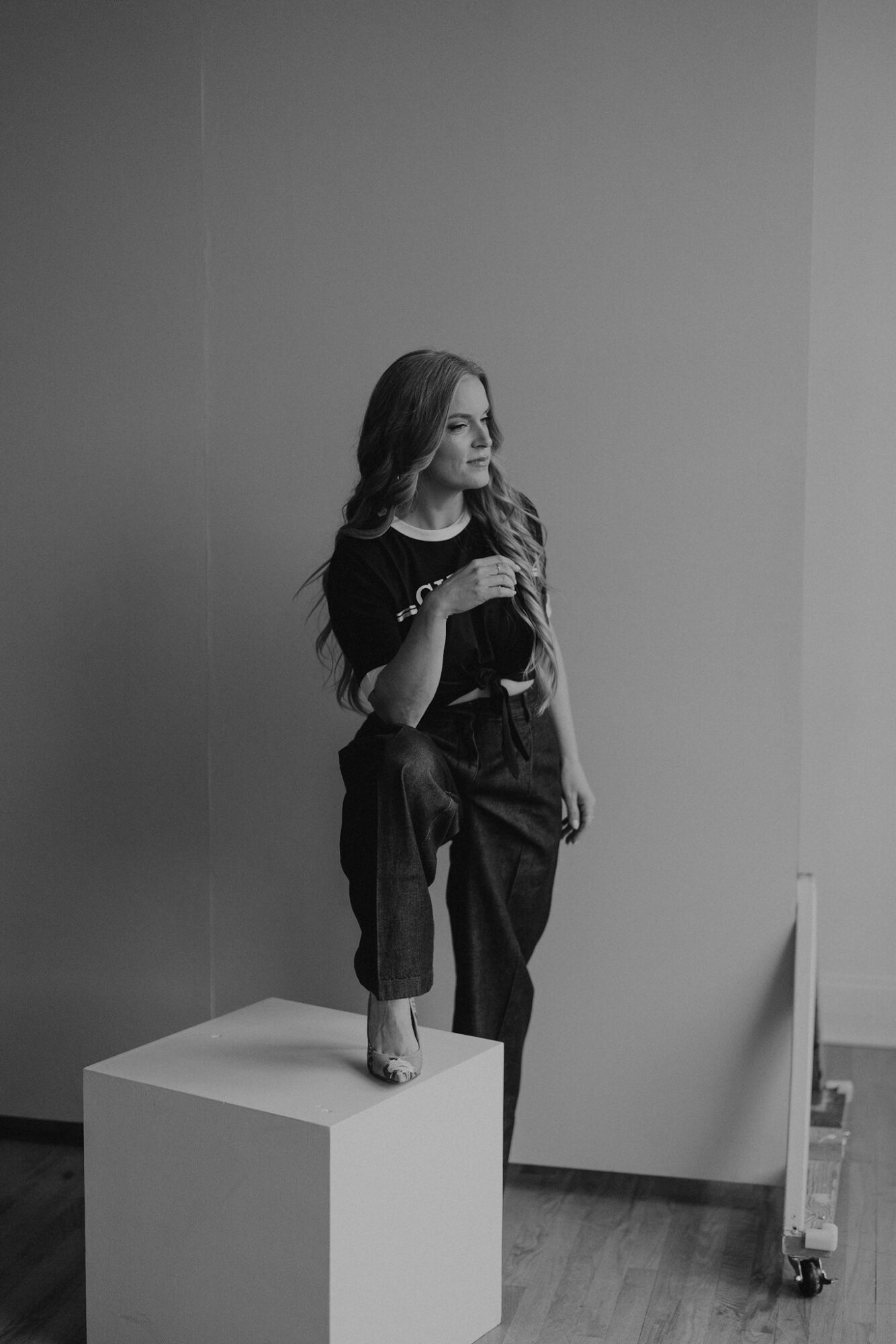 Woman standing on a pedestal in a room, holding a phone, with plain walls and a wooden floor.