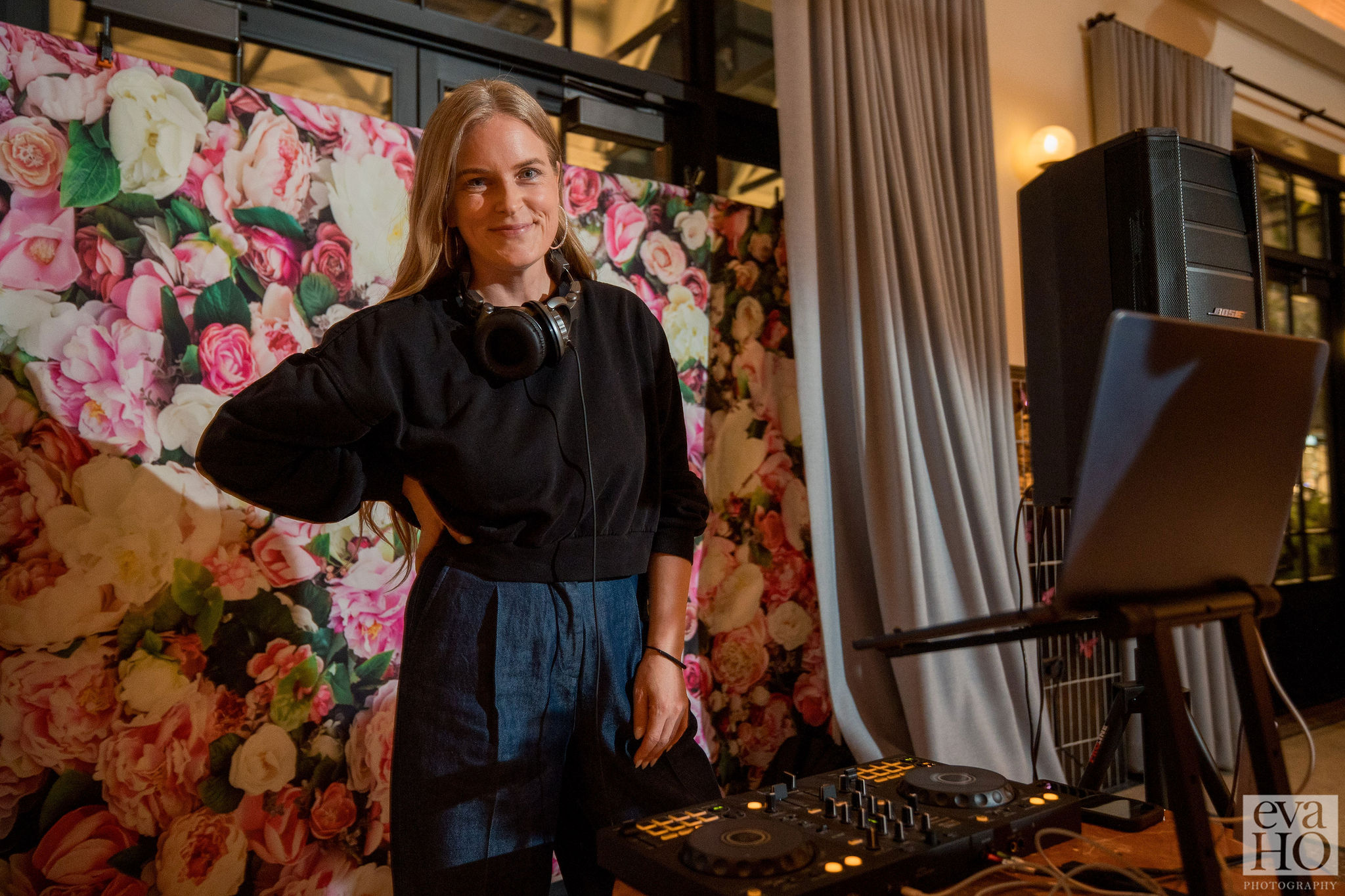 Woman standing in front of a floral backdrop, wearing a black top and blue pants, with DJ equipment and a laptop nearby.