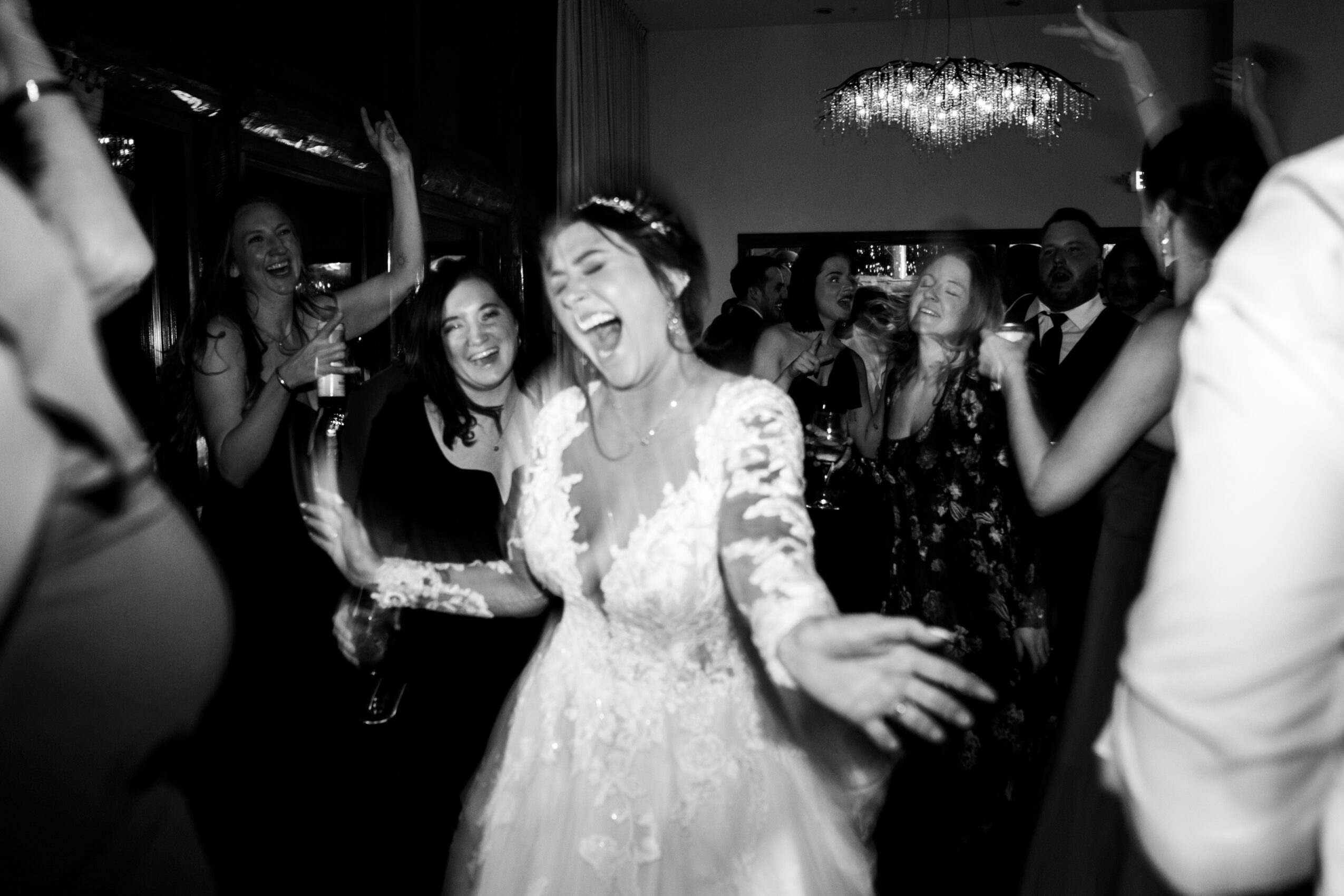 Group of people dancing and laughing at a celebration, woman in wedding dress in center, chandelier overhead, black and white photo.