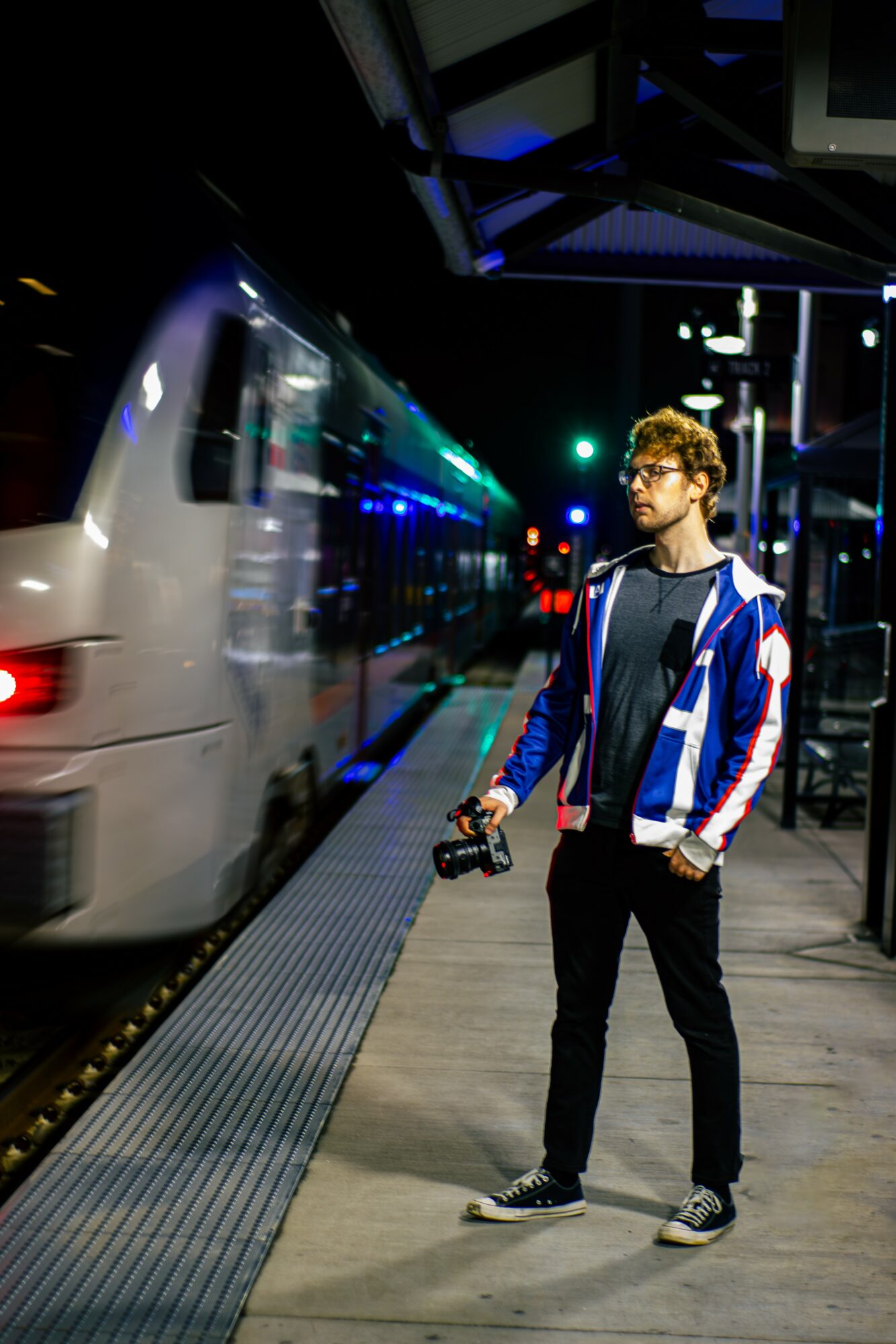 Young man with glasses and a backpack stands on a train platform at night, holding a camera, with a moving train behind him.