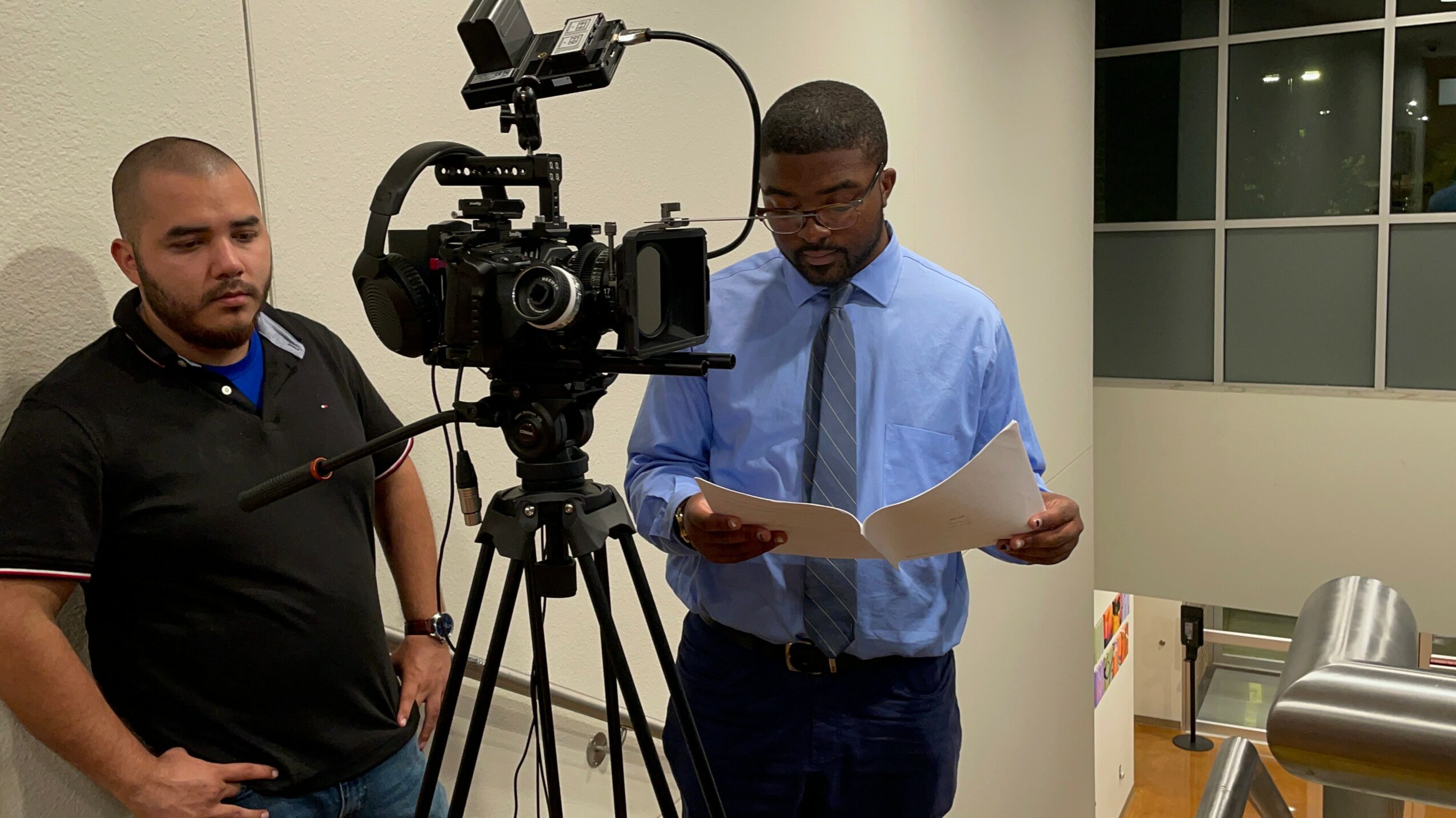 Two men in a room with a camera on a tripod, one reading a document, the other observing.