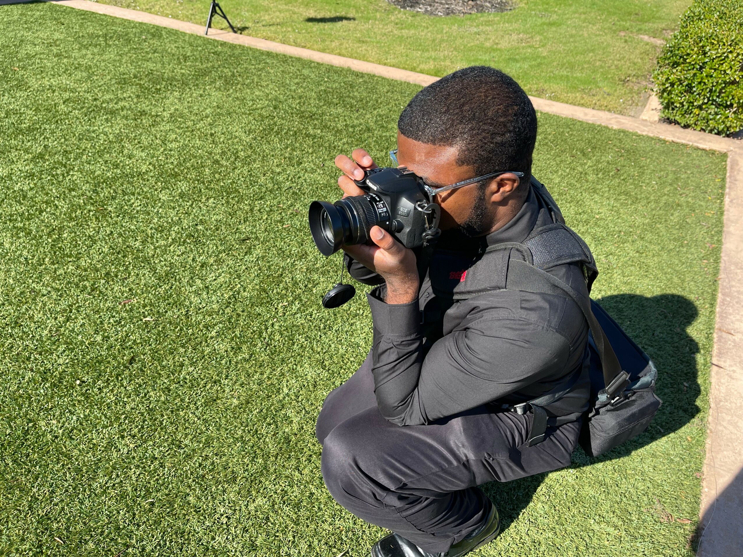 Person crouching on grass, taking a photo with a camera, outdoors on a sunny day.