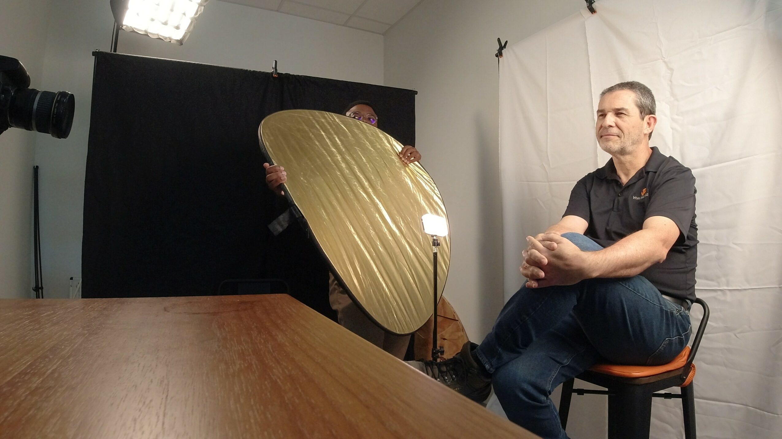 Person sitting on a chair in a photography studio, with a reflector and a black backdrop behind them.
