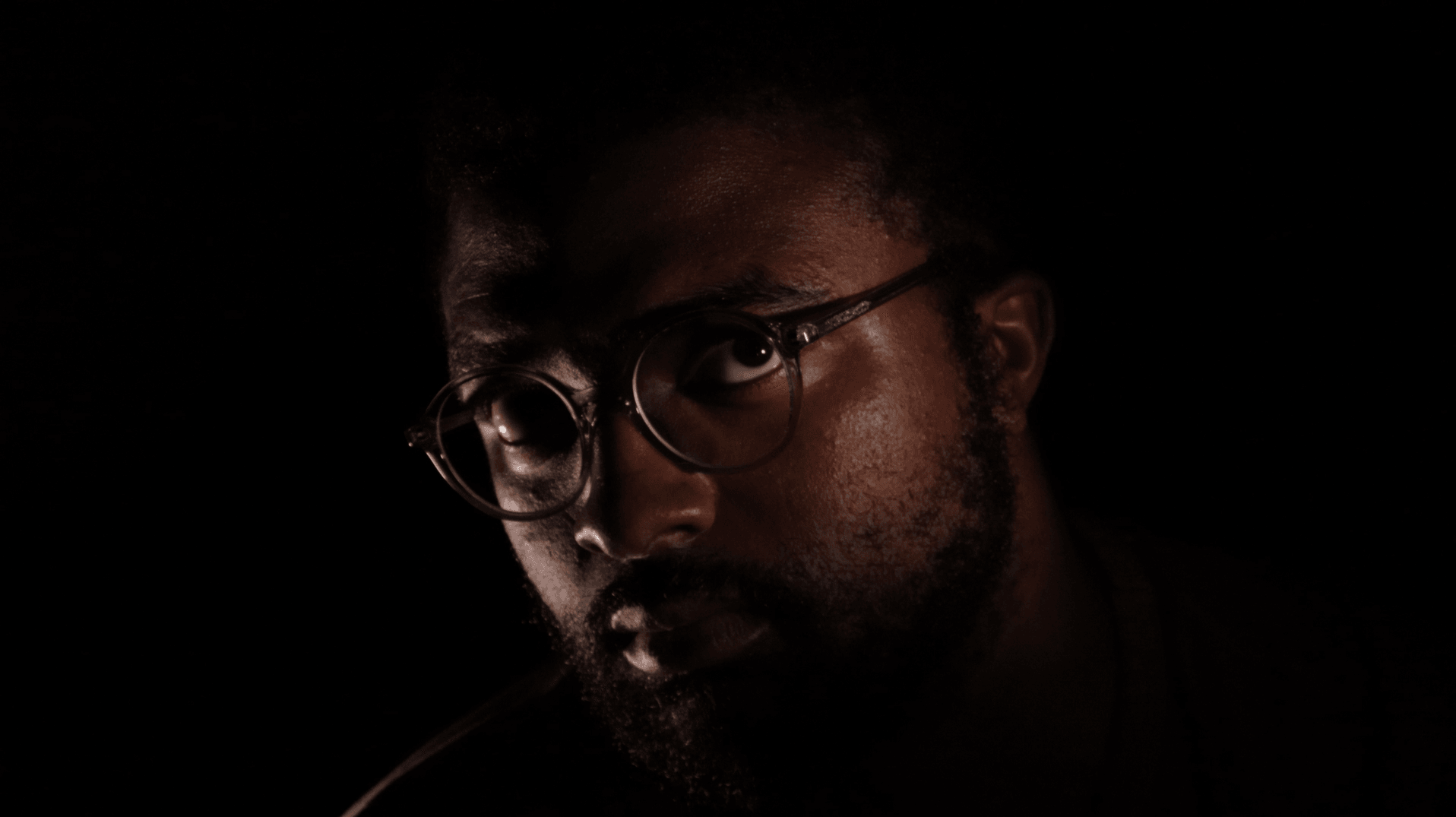 Portrait of a person with glasses, dark background, low lighting highlighting face features.