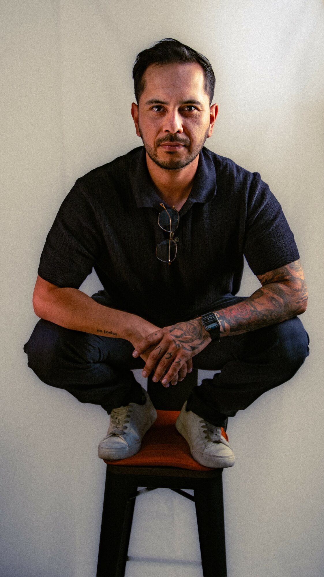 Man crouching on a chair, wearing a black shirt, glasses hanging from his collar, with tattoos on his left arm, against a plain background.
