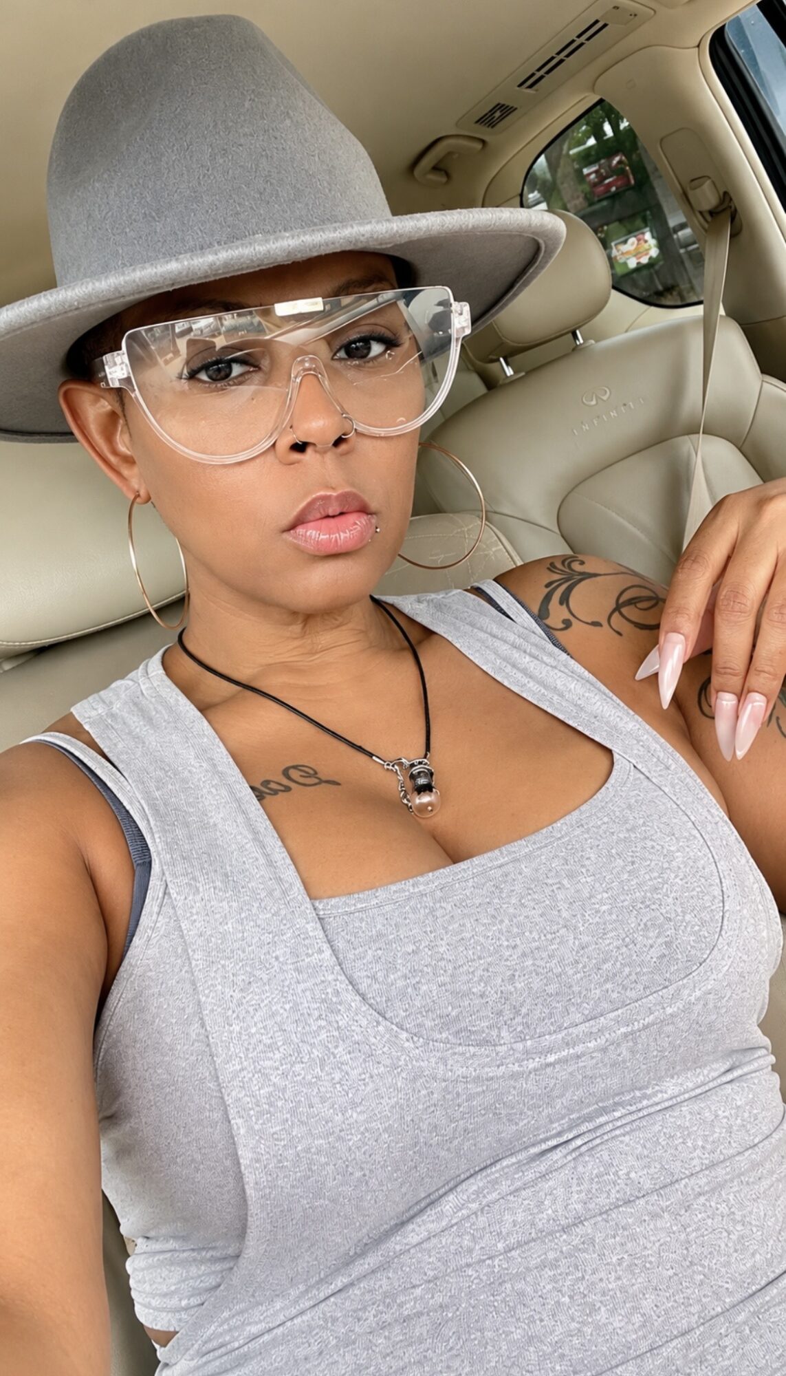 Woman wearing glasses and a wide-brimmed hat, sitting in a car, looking at the camera, with tattoos visible on her arm.
