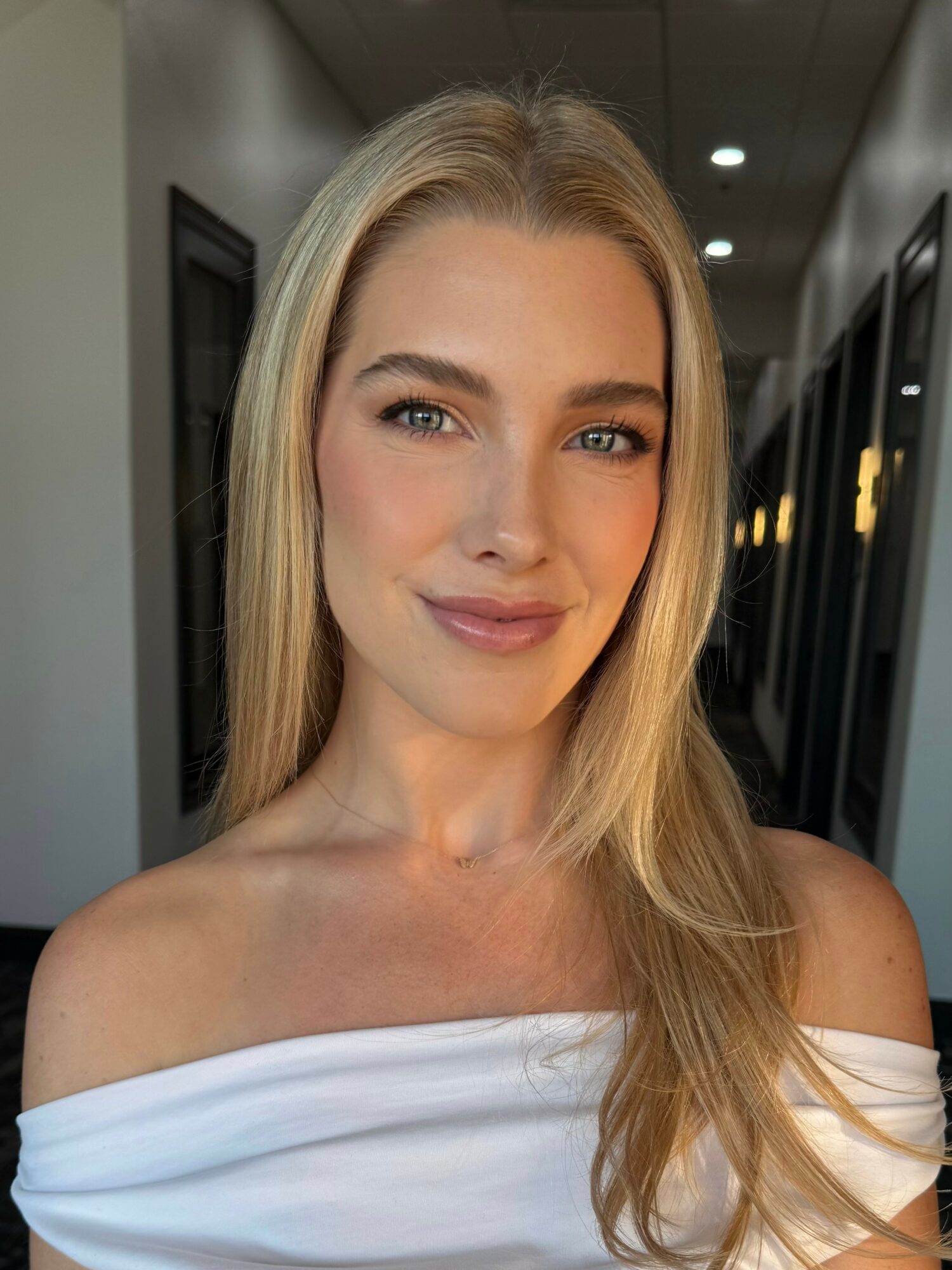 Young woman with long blonde hair smiling, wearing an off-shoulder white top, in a hallway with glass doors.