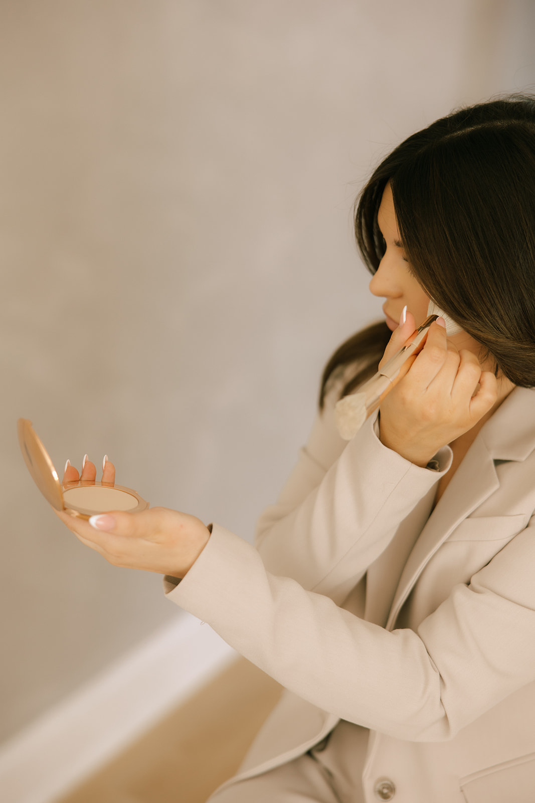 Woman with dark hair looking at her phone, touching her face with her right hand, wearing a light-colored blazer.
