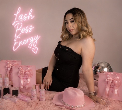 Woman in black dress standing at a table with pink gifts, hat, and a neon sign reading 'Fash Boss Energy' with a heart.