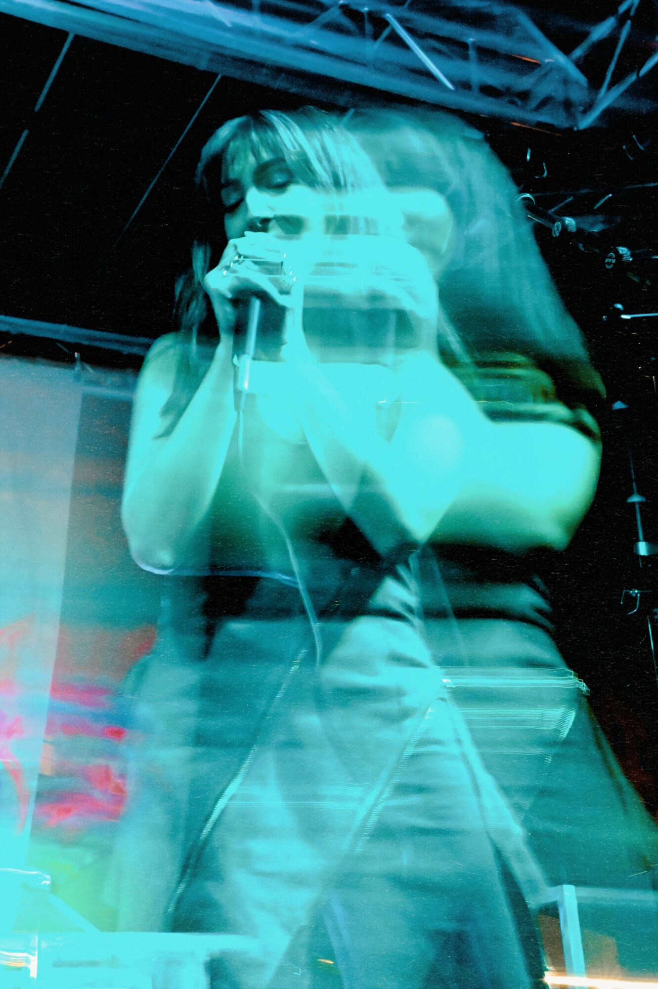 A person with long hair holding a microphone, performing on stage with a dark background and stage lighting.
