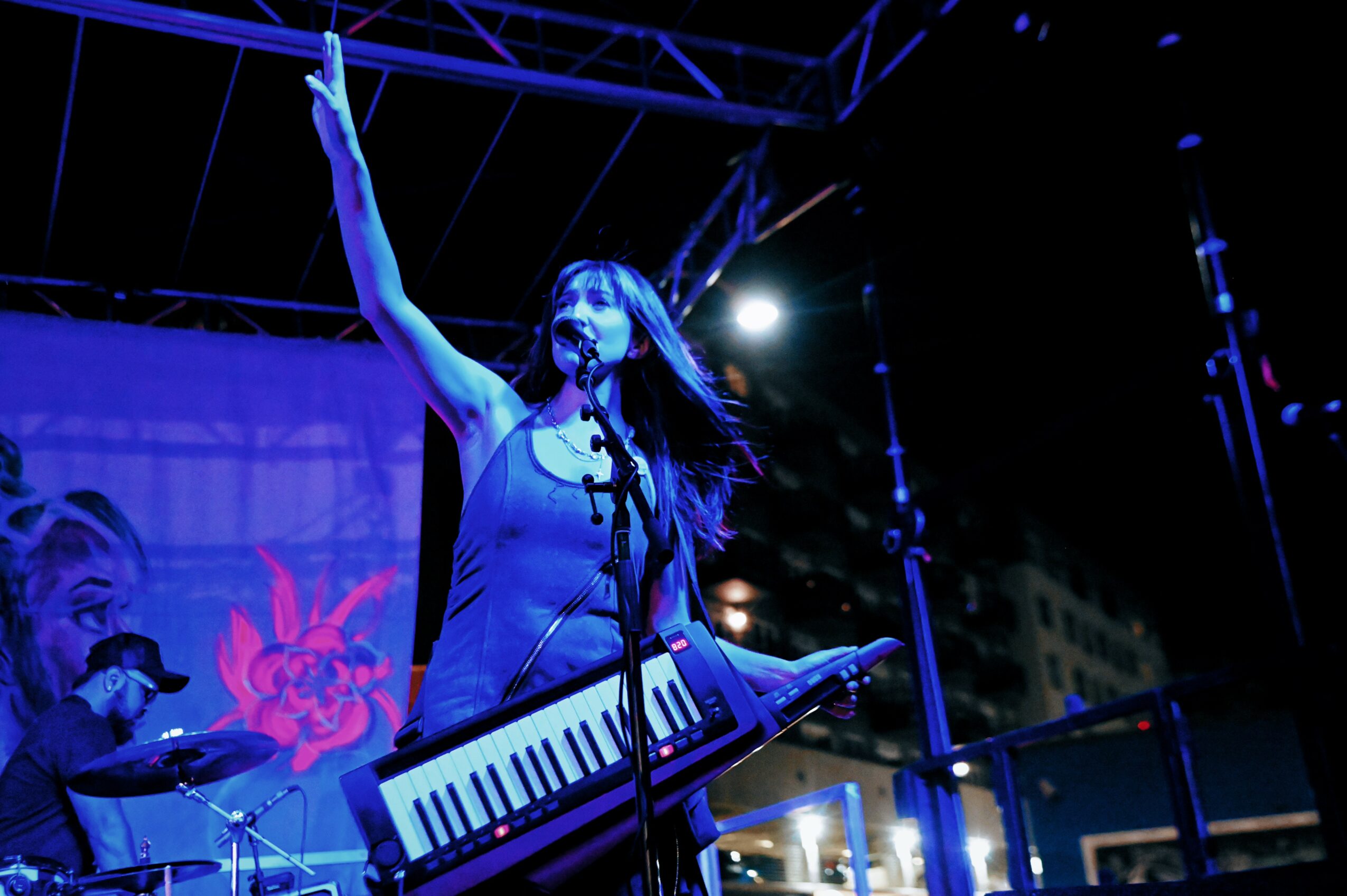 Female musician singing into a microphone, playing a keytar, with her arm raised on stage at night.