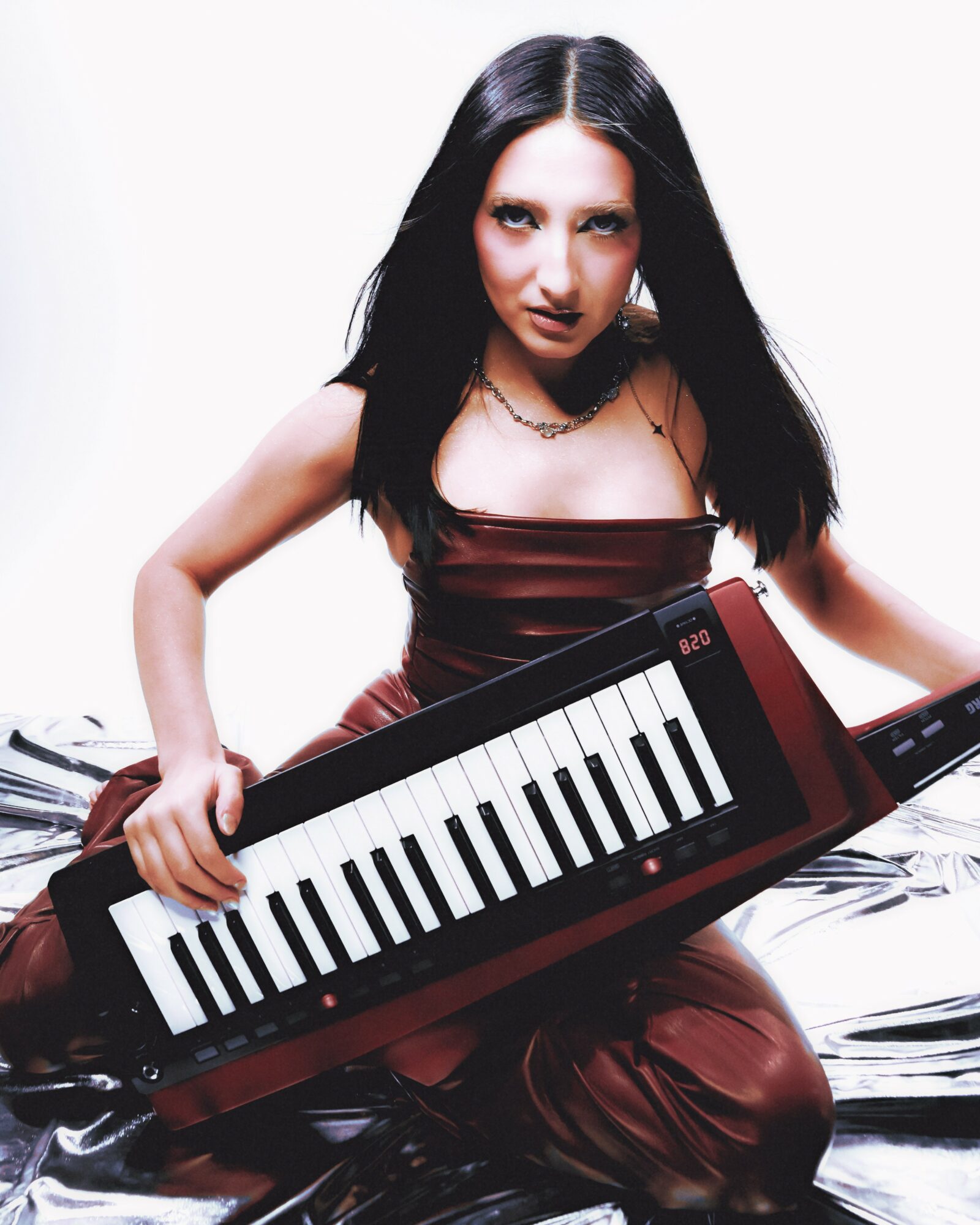 Woman with long dark hair holding a keyboard, sitting on a reflective surface, wearing a dark dress, looking at the camera.