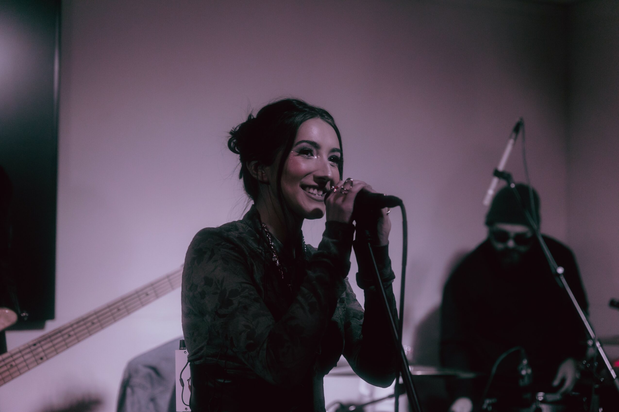Woman singing into a microphone on stage with a person in the background, dim lighting, smiling face, dark clothing.