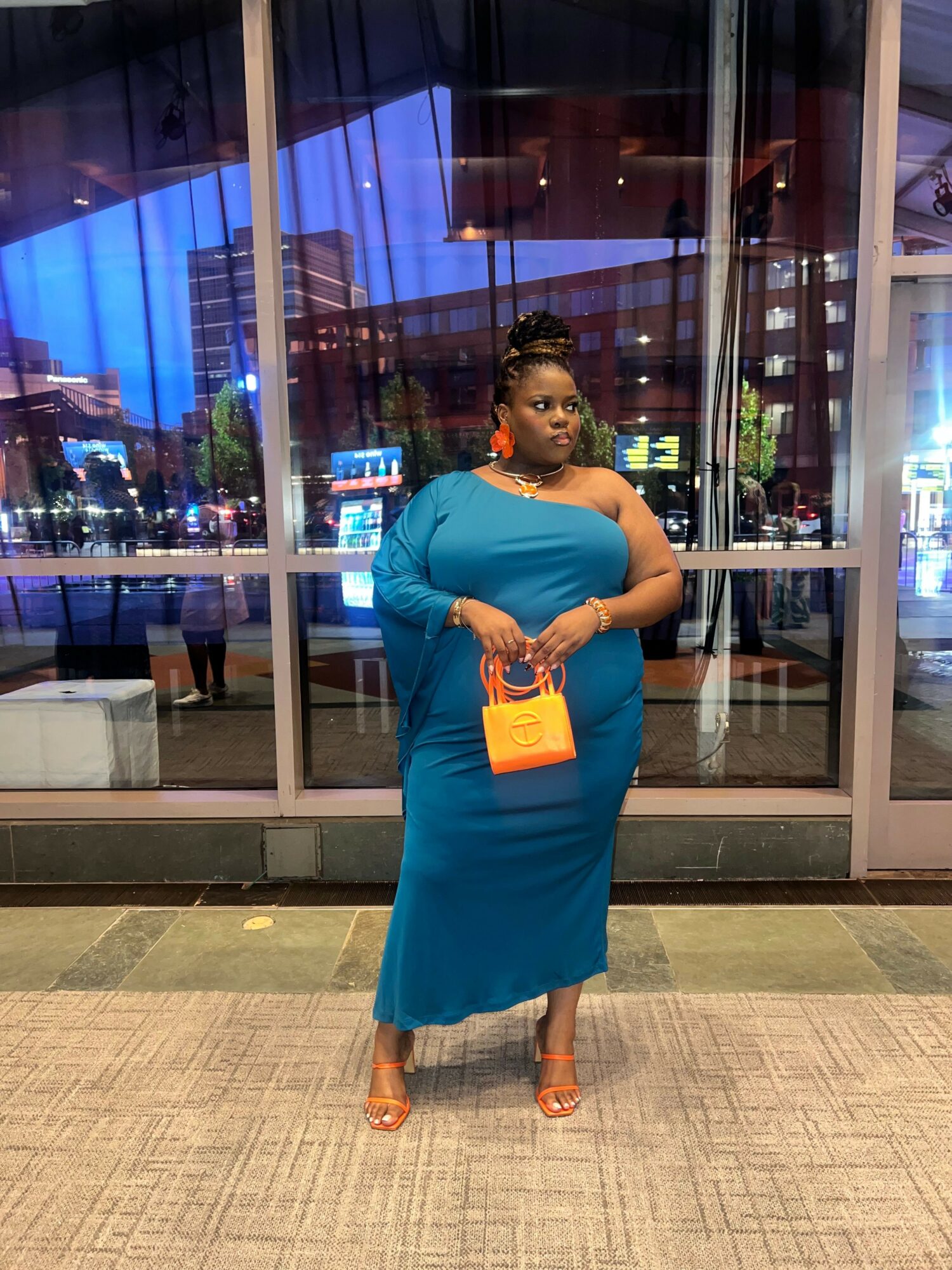 Woman in blue dress holding a yellow clutch bag, standing indoors near glass doors, with city lights visible outside.