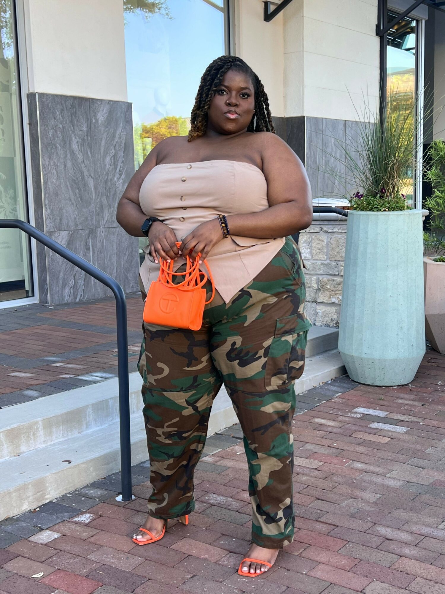 Woman standing outdoors holding an orange bag, wearing a beige top, camouflage pants, and sandals.