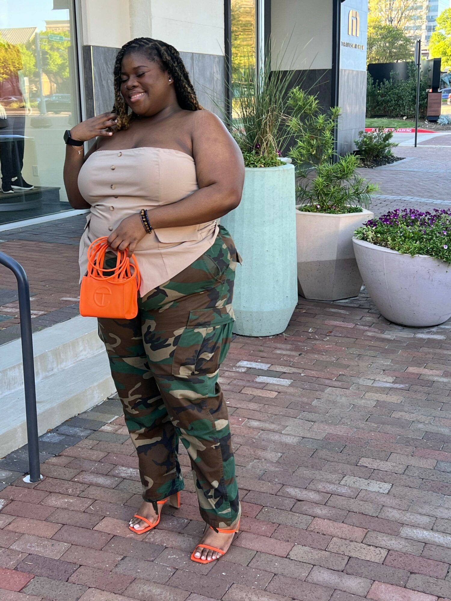 Woman with long dark hair, wearing camouflage pants and orange sandals, holding a beige bag, standing on brick sidewalk.