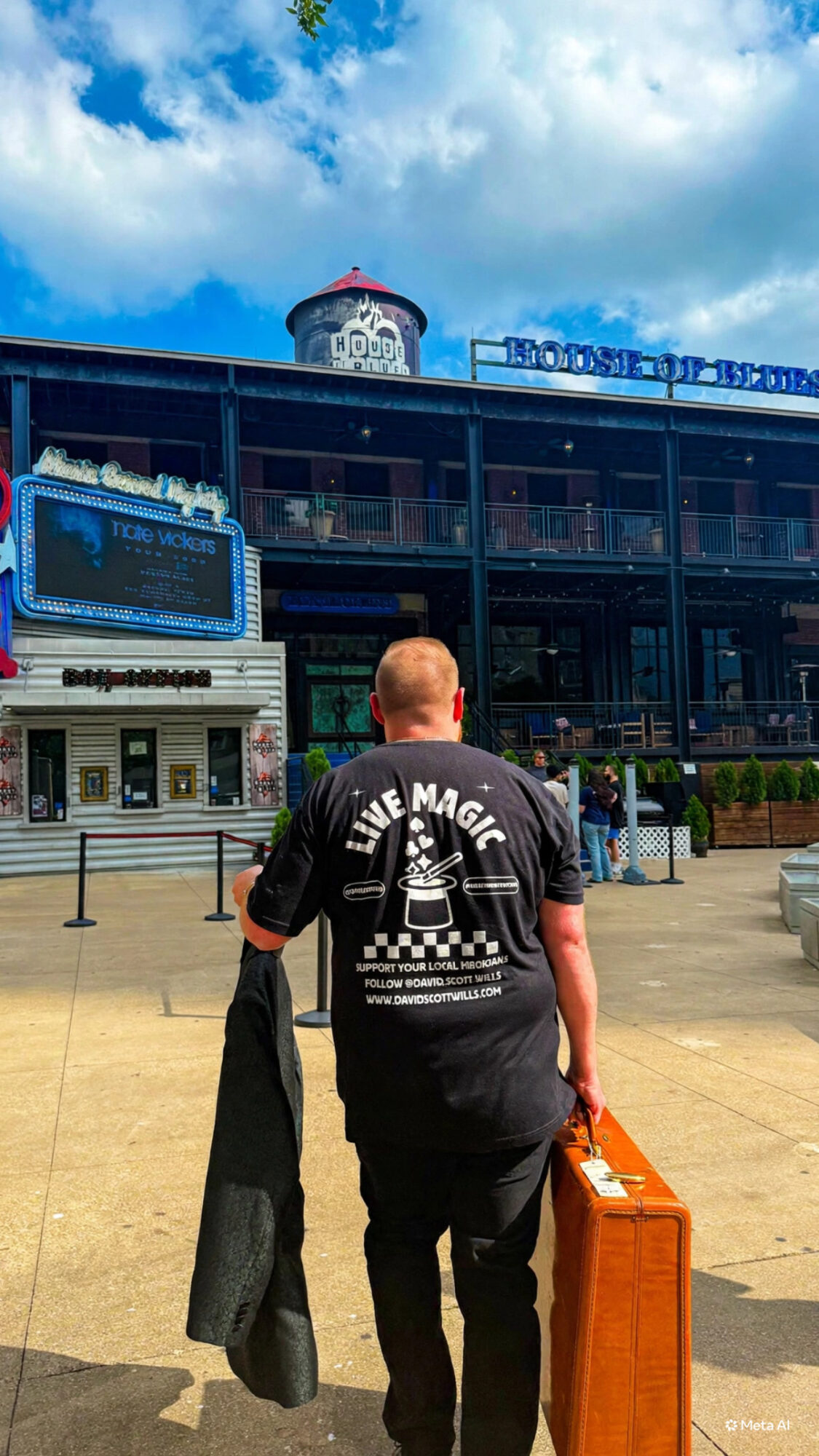 Person walking away from camera in front of a large building with a sign, carrying a jacket and a suitcase.