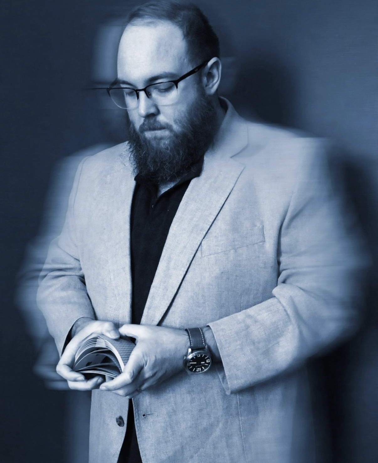 Man with glasses and beard in a light blazer looks at a book, with a blurred background.
