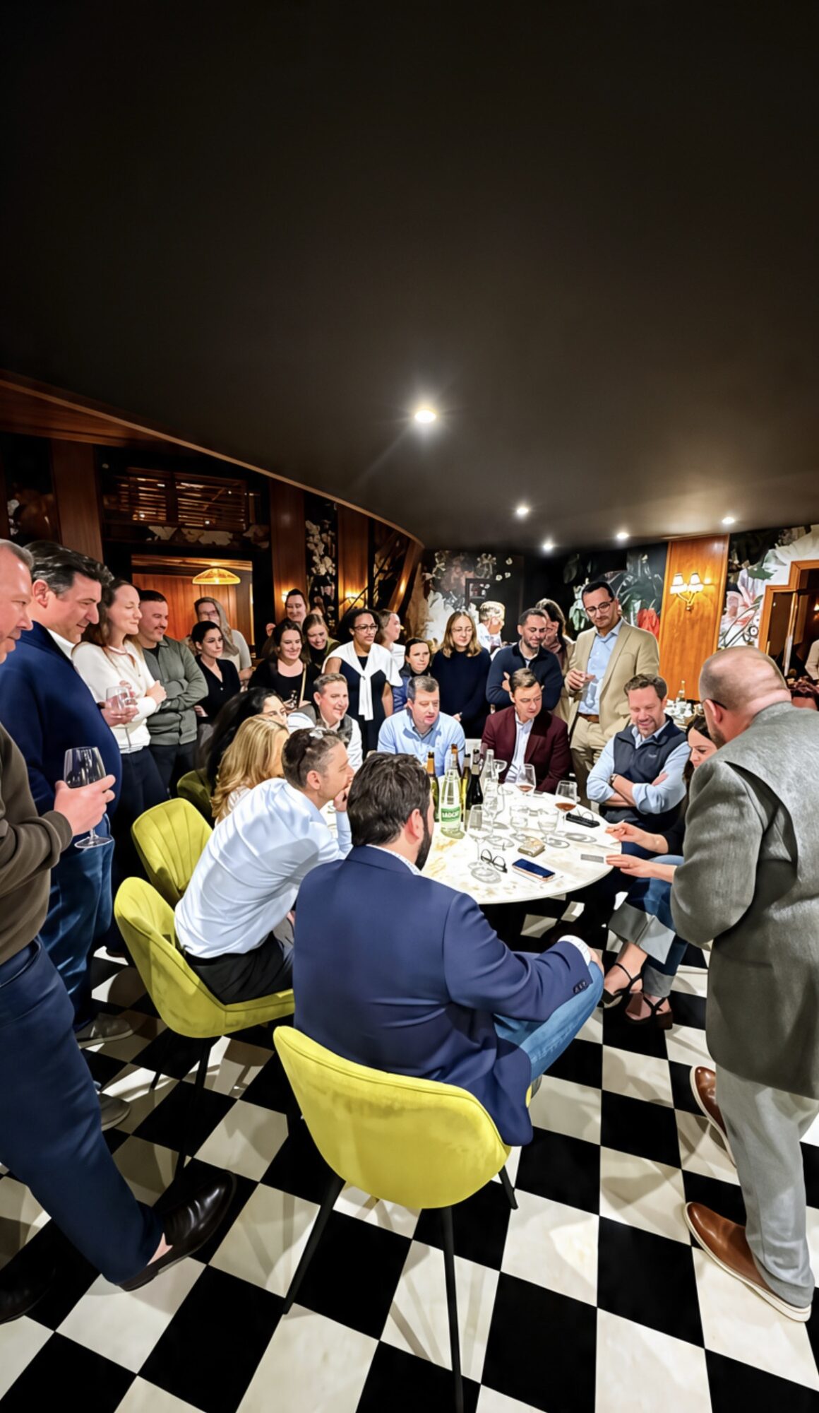 Group of people gathered around a table in a restaurant, some standing and some seated, with a checkered floor and warm lighting.