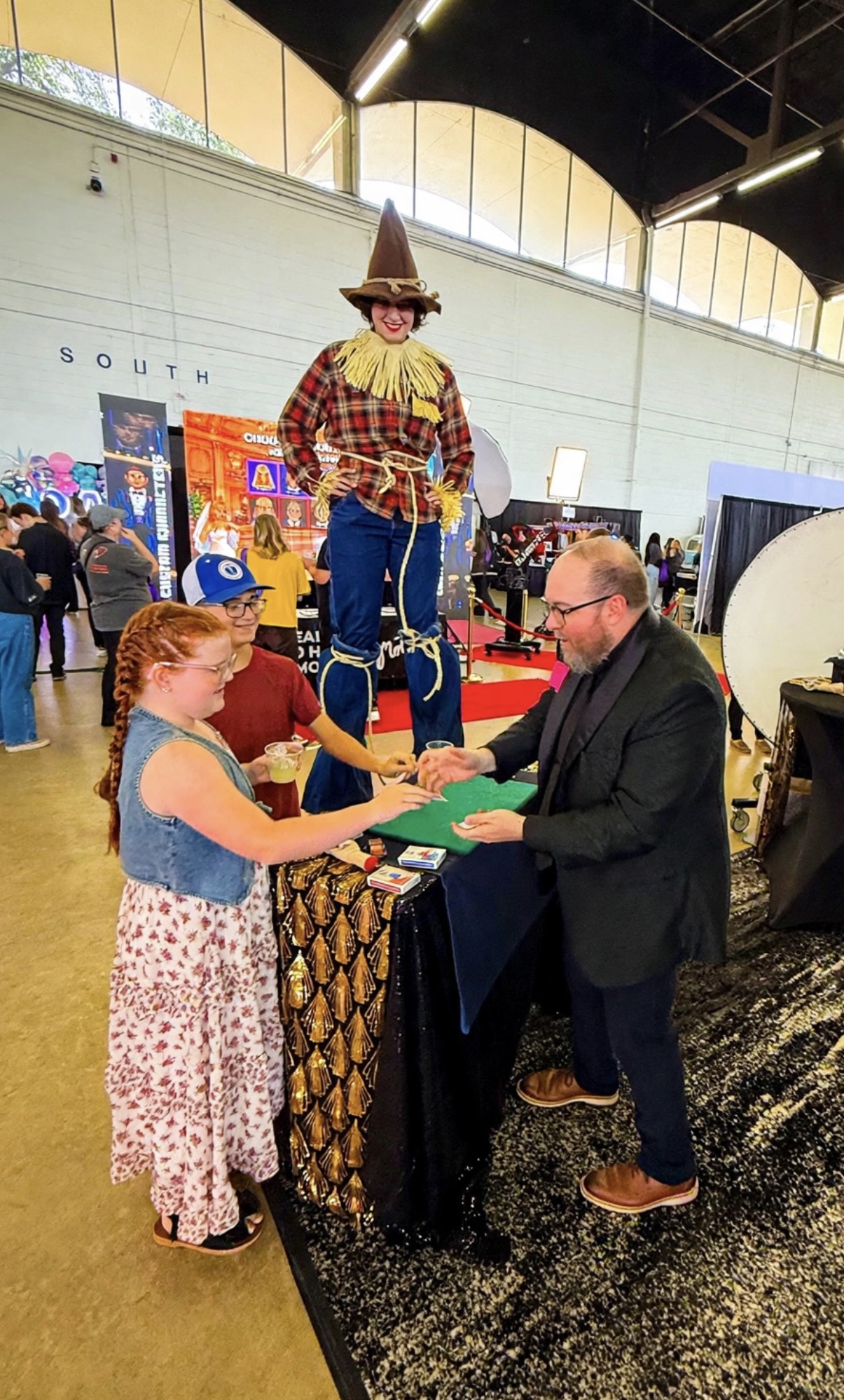 Child receiving an award from a man at a booth, with a clown figure in the background, in an indoor event space.