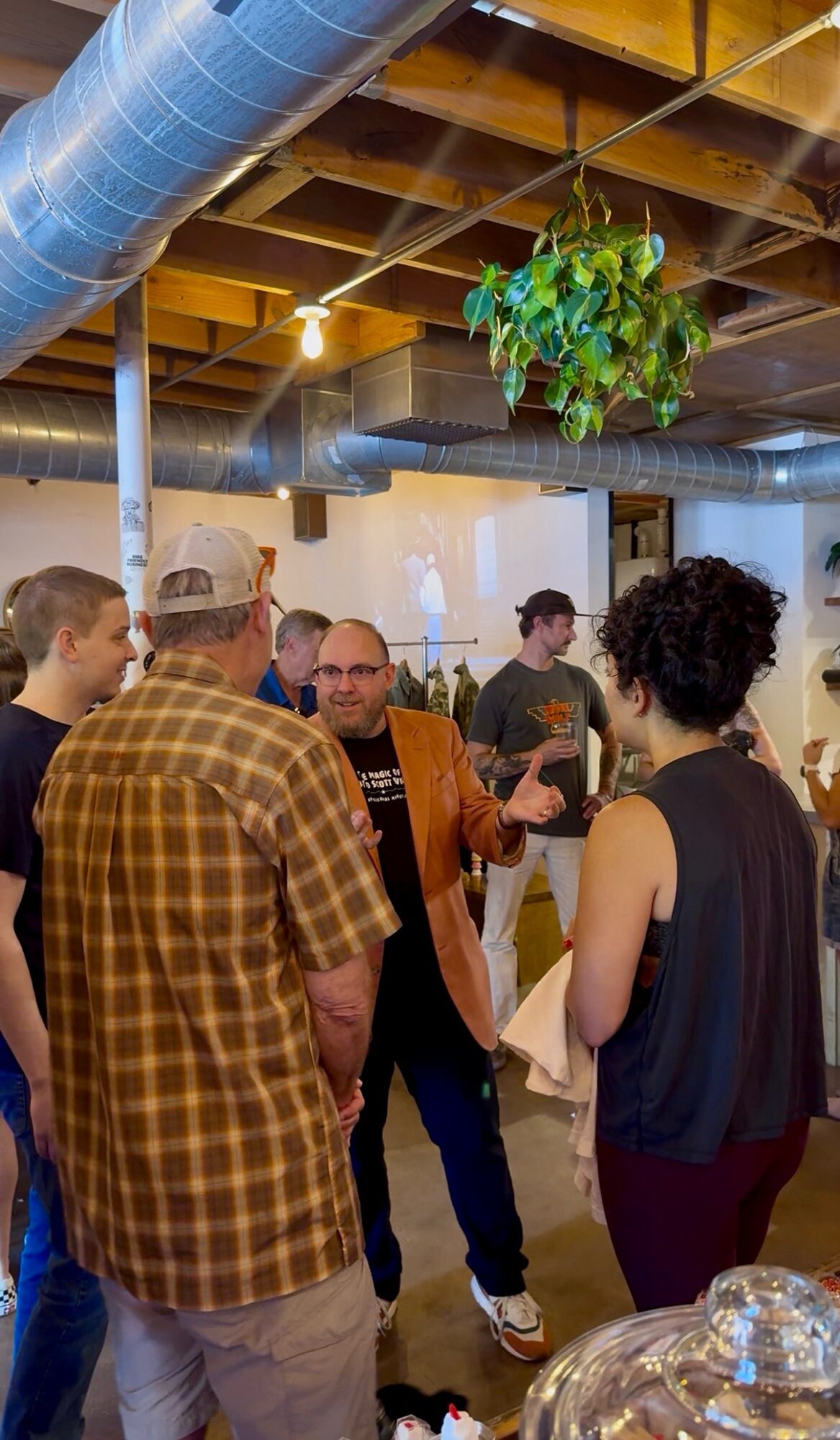 Group of people socializing indoors with exposed ceiling beams and hanging plant, some holding drinks, engaged in conversation.
