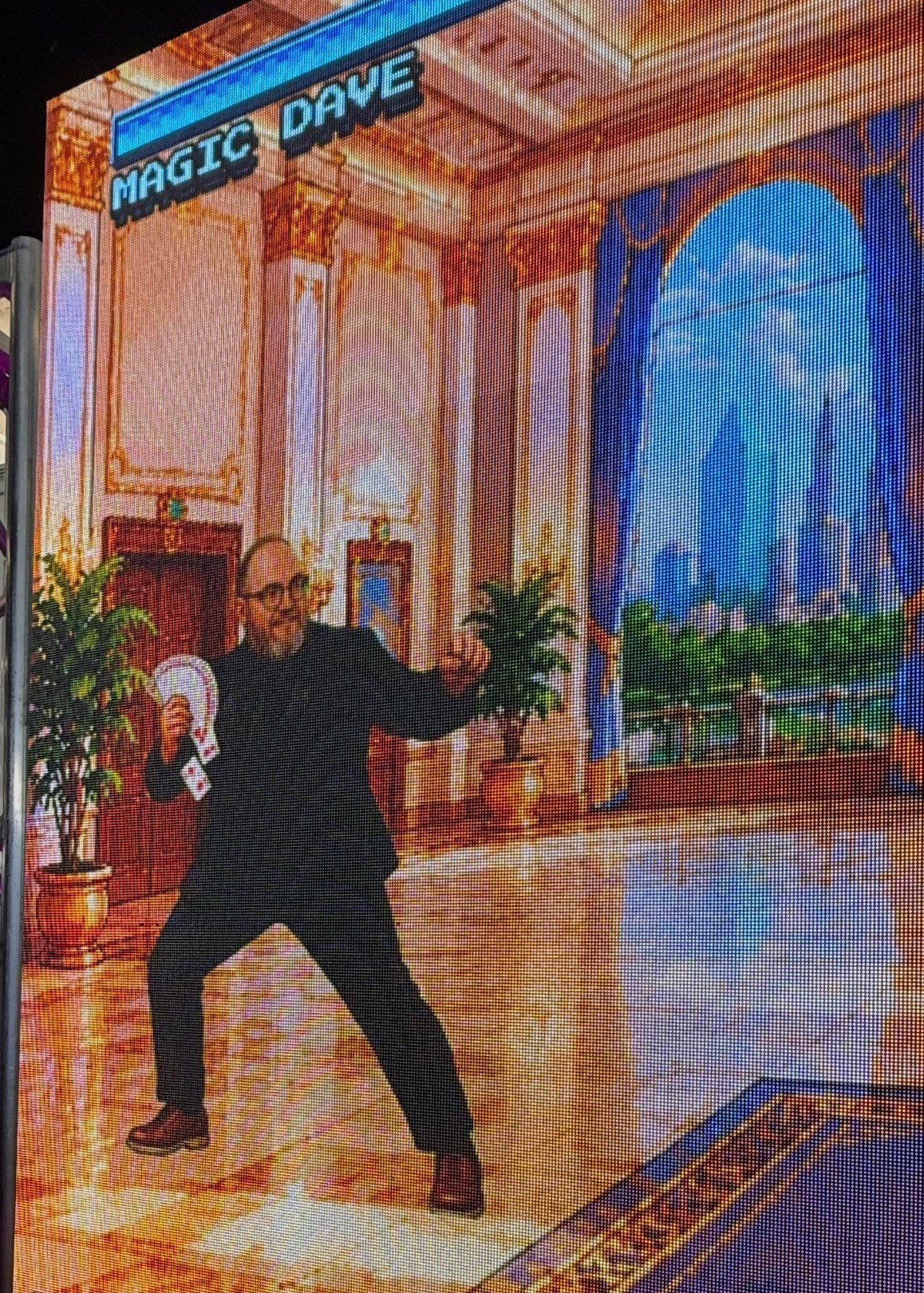 Person in black suit performing magic trick in ornate room with large window and cityscape view, holding props, smiling.