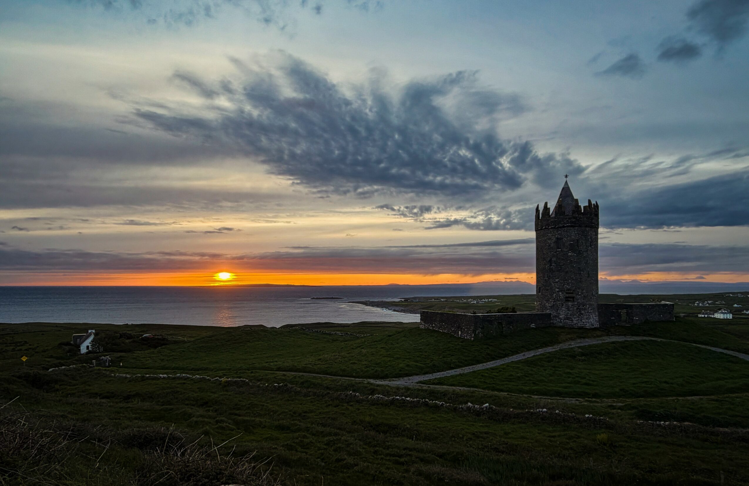 Sunset over a landscape with a castle tower and a body of water, cloudy sky, and grassy foreground.