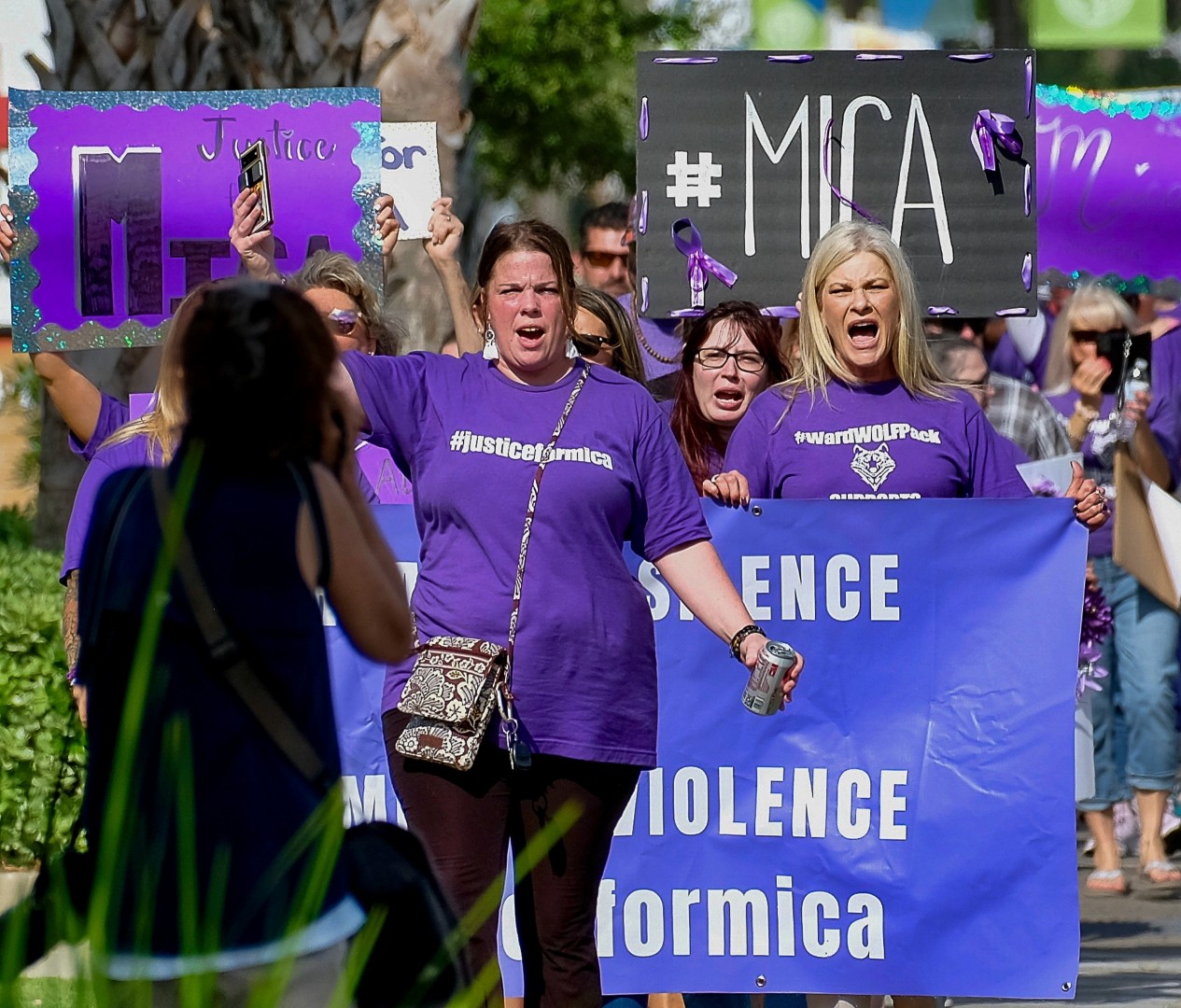 Group of women protesting, holding signs and banners, some wearing purple shirts, outdoors on a sunny day.