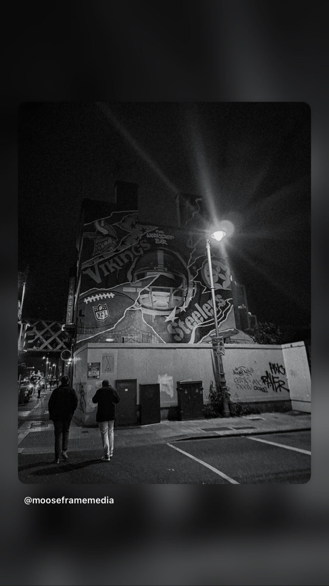 Two people walk past a mural on a building at night, illuminated by a streetlight, with a dark sky background.