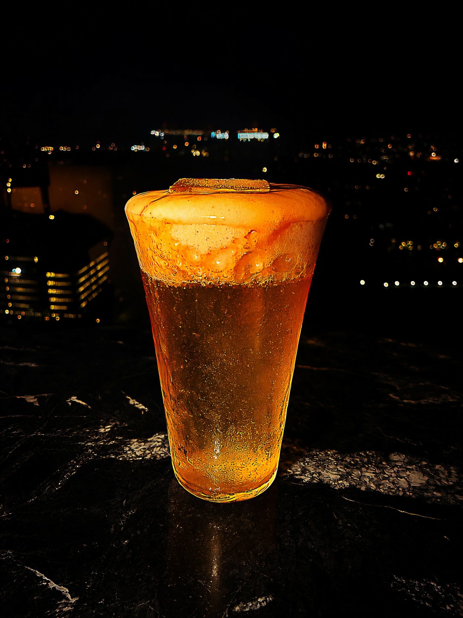 A glass of beer with foam on top, set on a dark surface against a city nightscape background.