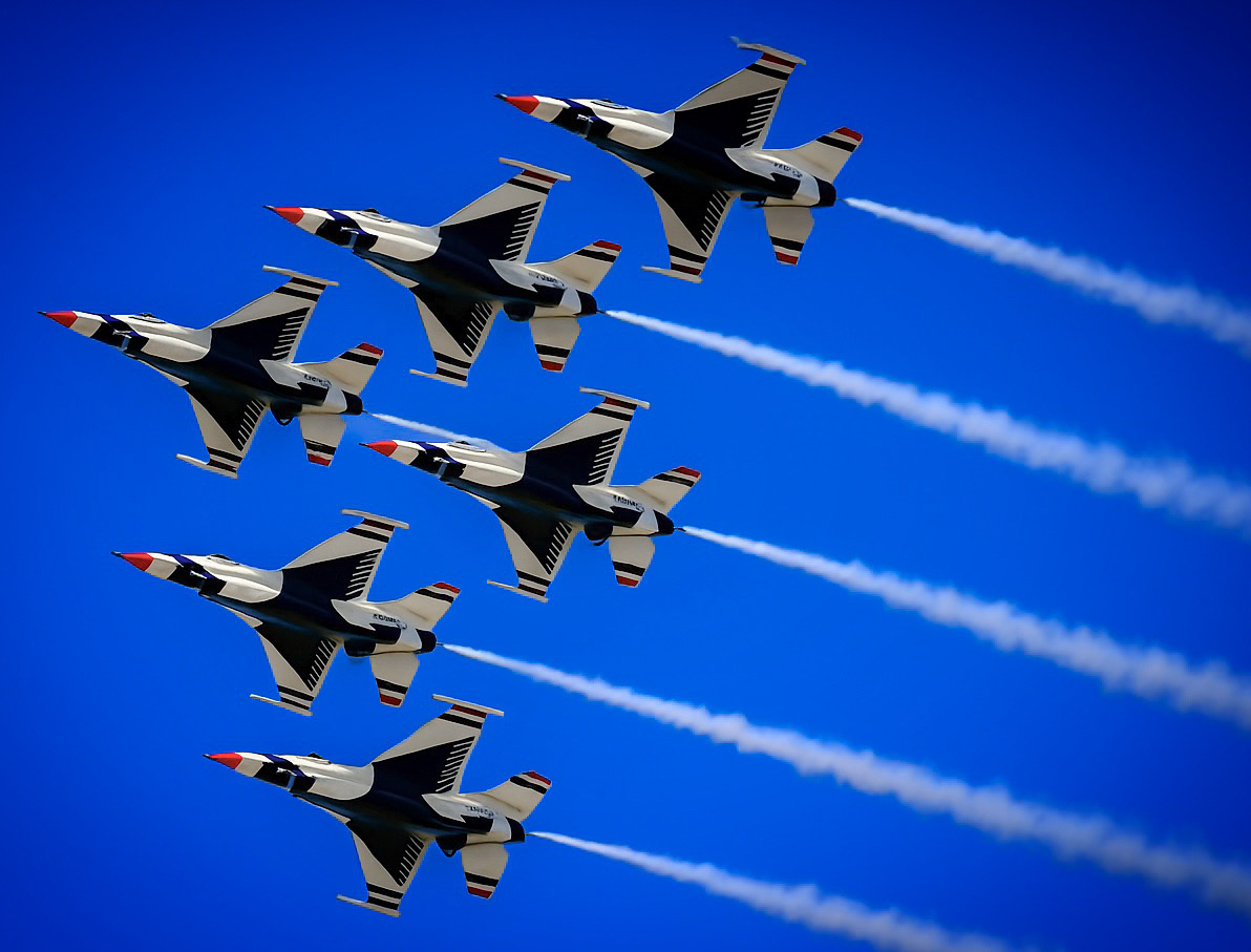 Six fighter jets flying in a tight formation against a blue sky, leaving white smoke trails behind them.