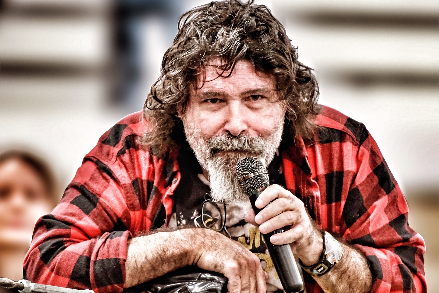 Man with curly hair and beard speaking into a microphone, wearing a red and black plaid shirt.