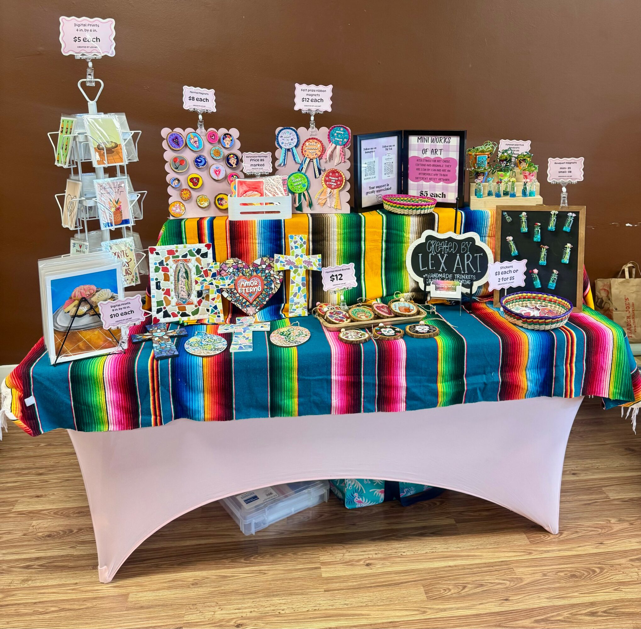 Display table with colorful Mexican textiles, jewelry, and art supplies against a brown wall, with signs and price tags.