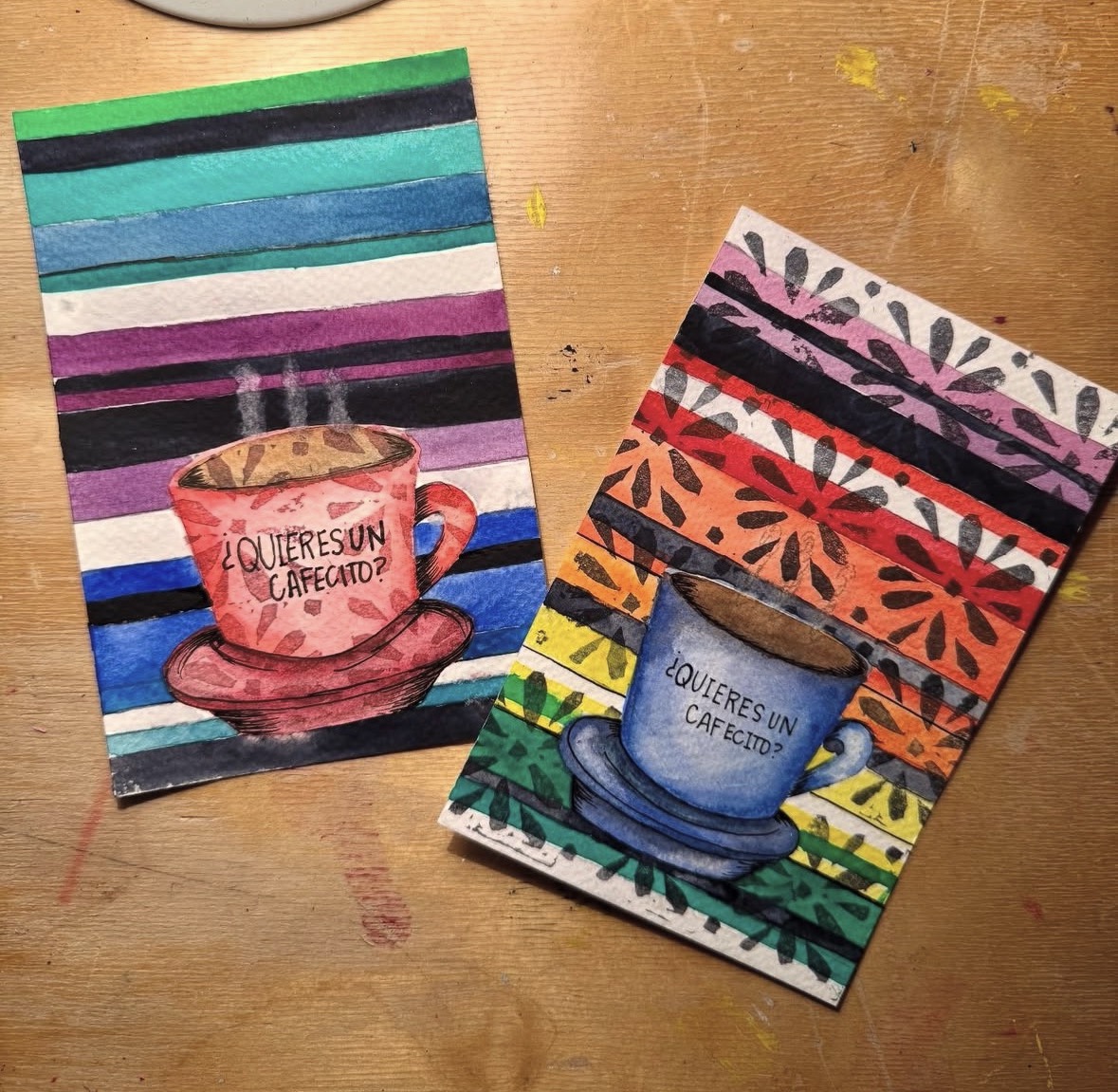 Two colorful paintings of coffee cups with Spanish text, one pink and one blue, on striped and floral backgrounds.