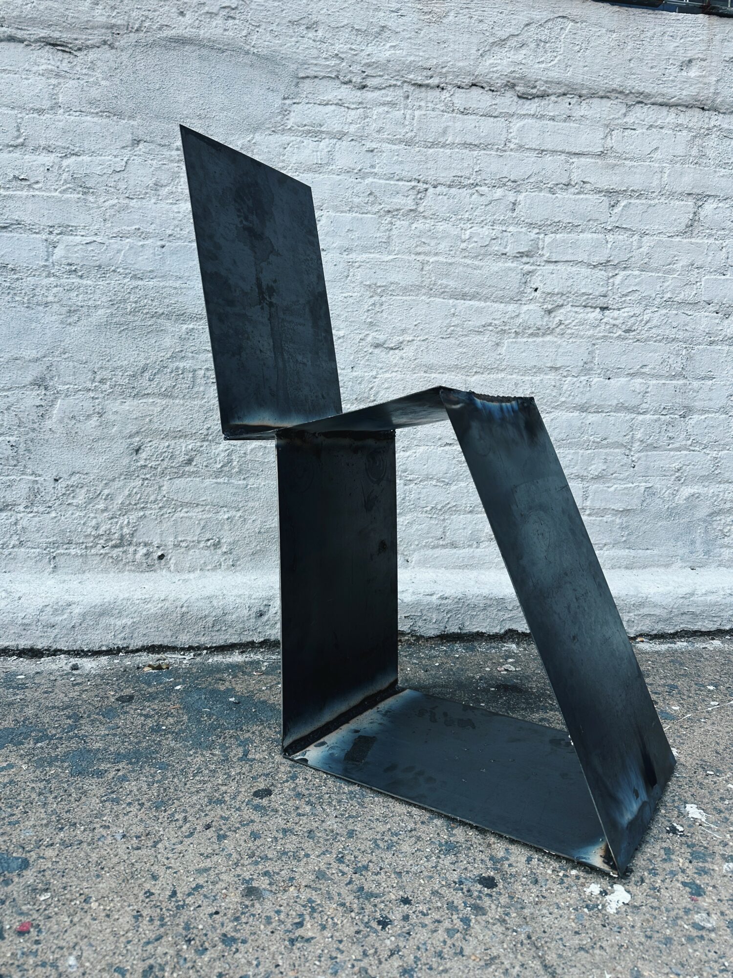 Metal chair with a modern, geometric design, placed outdoors against a white brick wall.