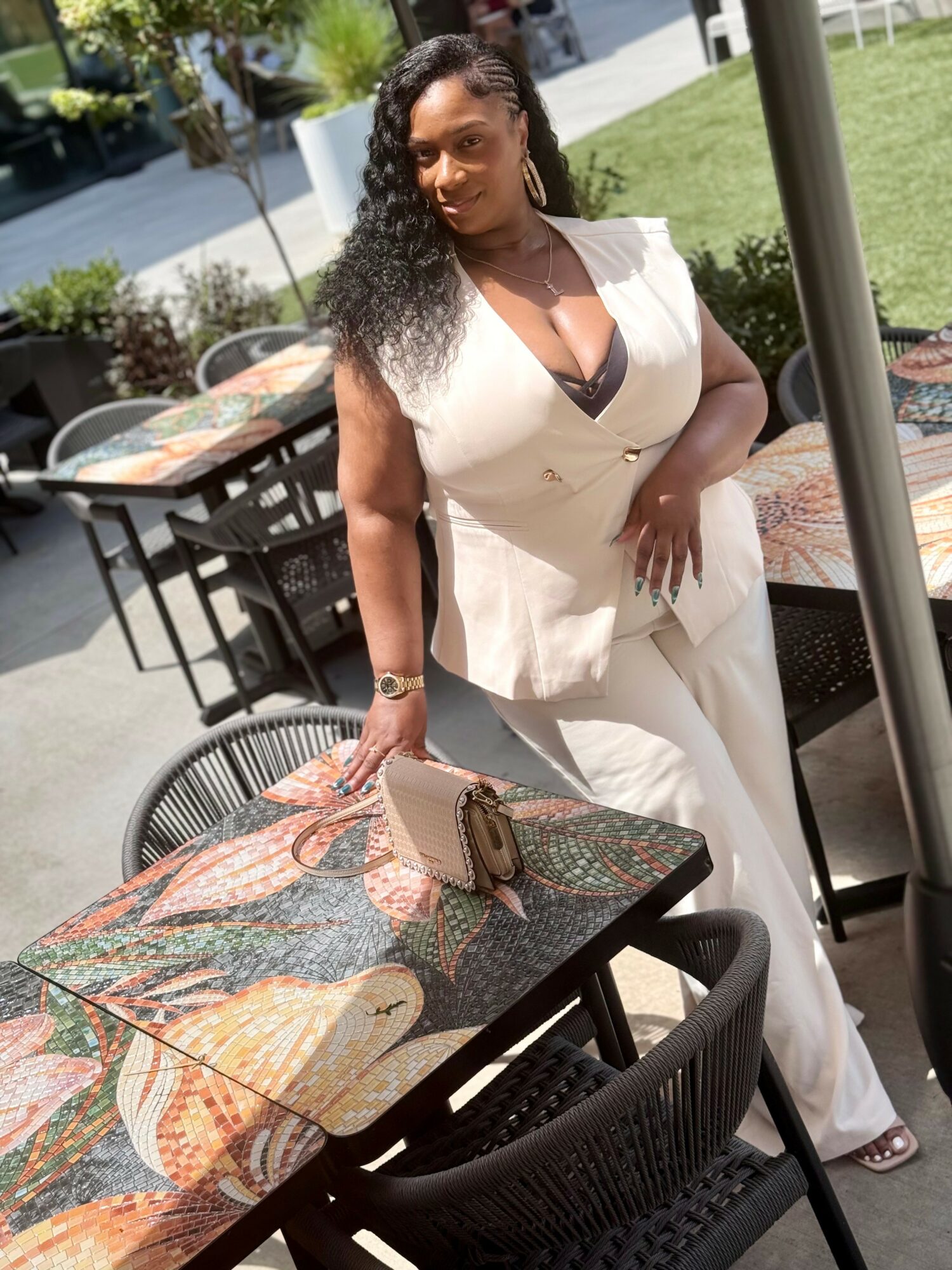 Woman in white outfit standing outdoors at a table with a colorful patterned surface, holding a purse, with greenery and outdoor furniture in background.