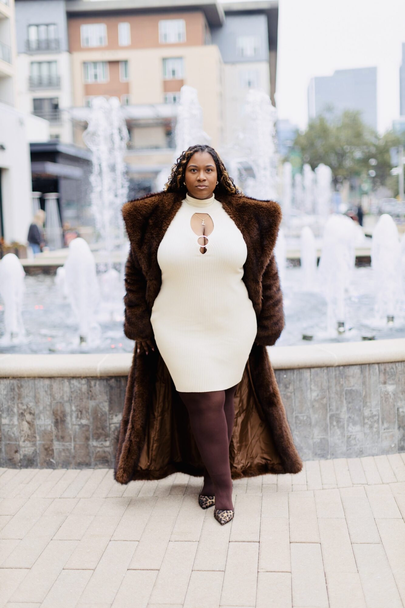 Woman in white dress and brown fur coat standing in front of a fountain in an urban setting.