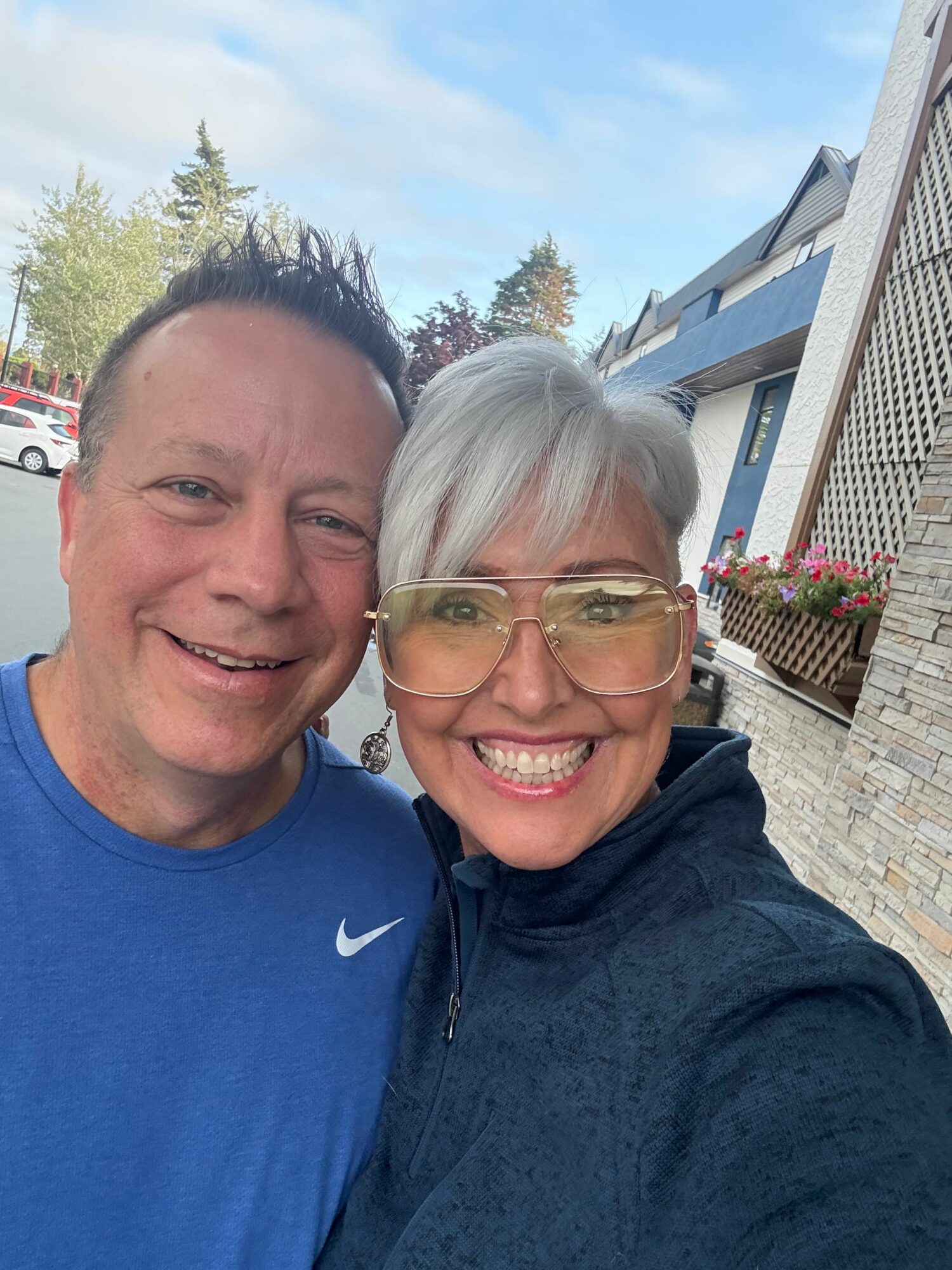 Two smiling people, one with gray hair and glasses, the other with dark hair, outdoors with buildings and trees in background.