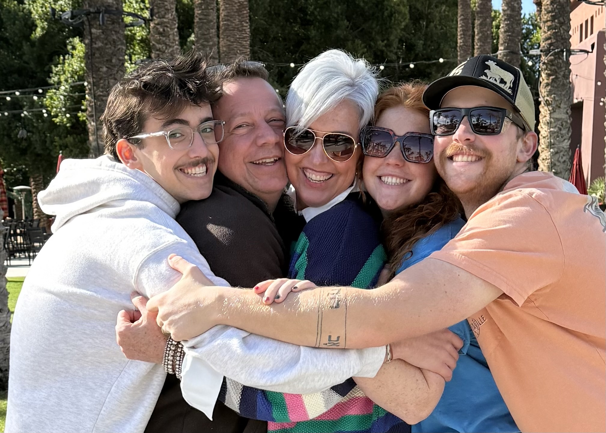 Group of five people hugging outdoors, smiling, with trees and string lights in background.