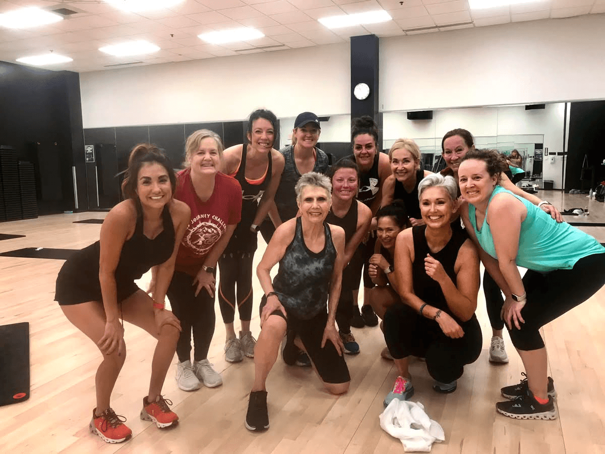 Group of ten women in workout clothes smiling in a fitness studio with mirrors and wooden floors.