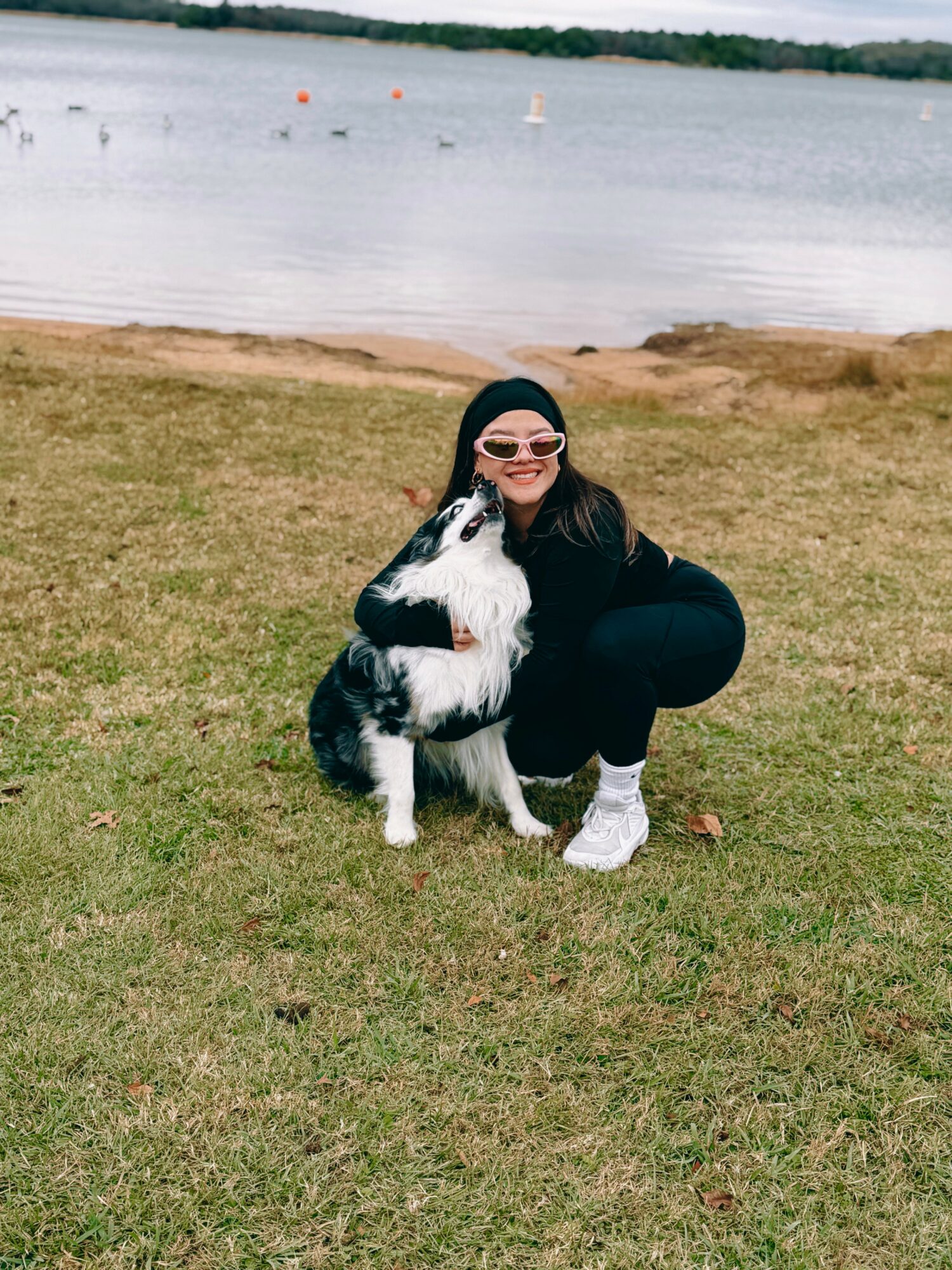 Person crouching next to a black and white dog on grass near water, wearing sunglasses and a black hoodie.