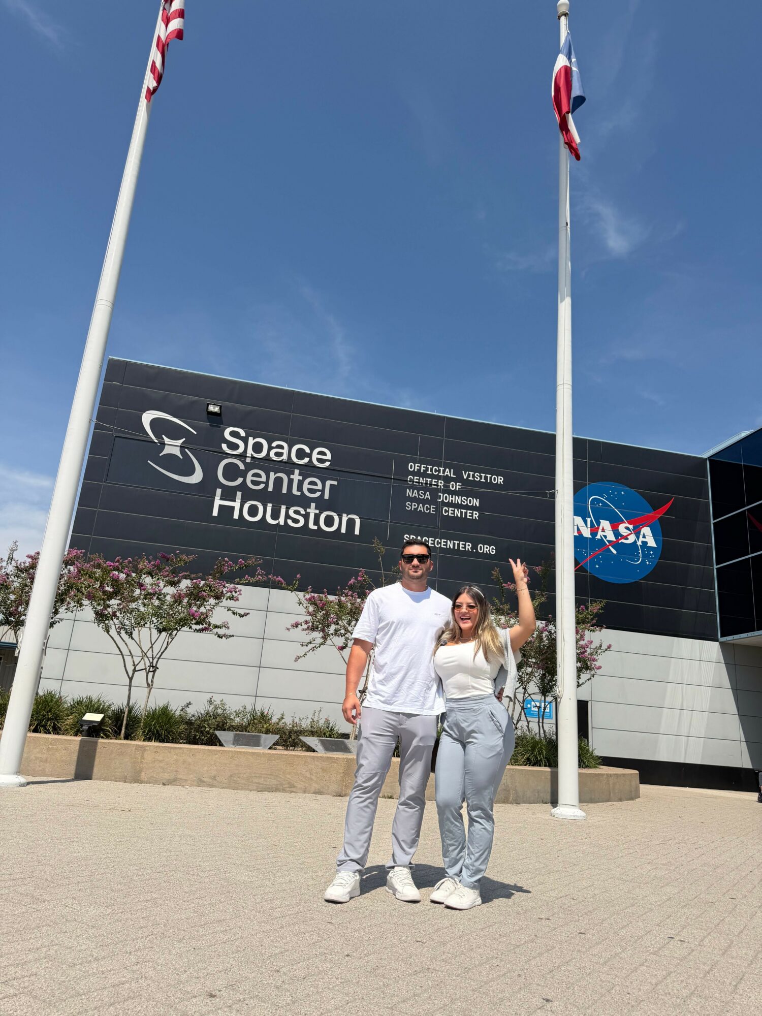 Two people stand outside Space Center Houston, with a sign and NASA logo behind them, under a blue sky.